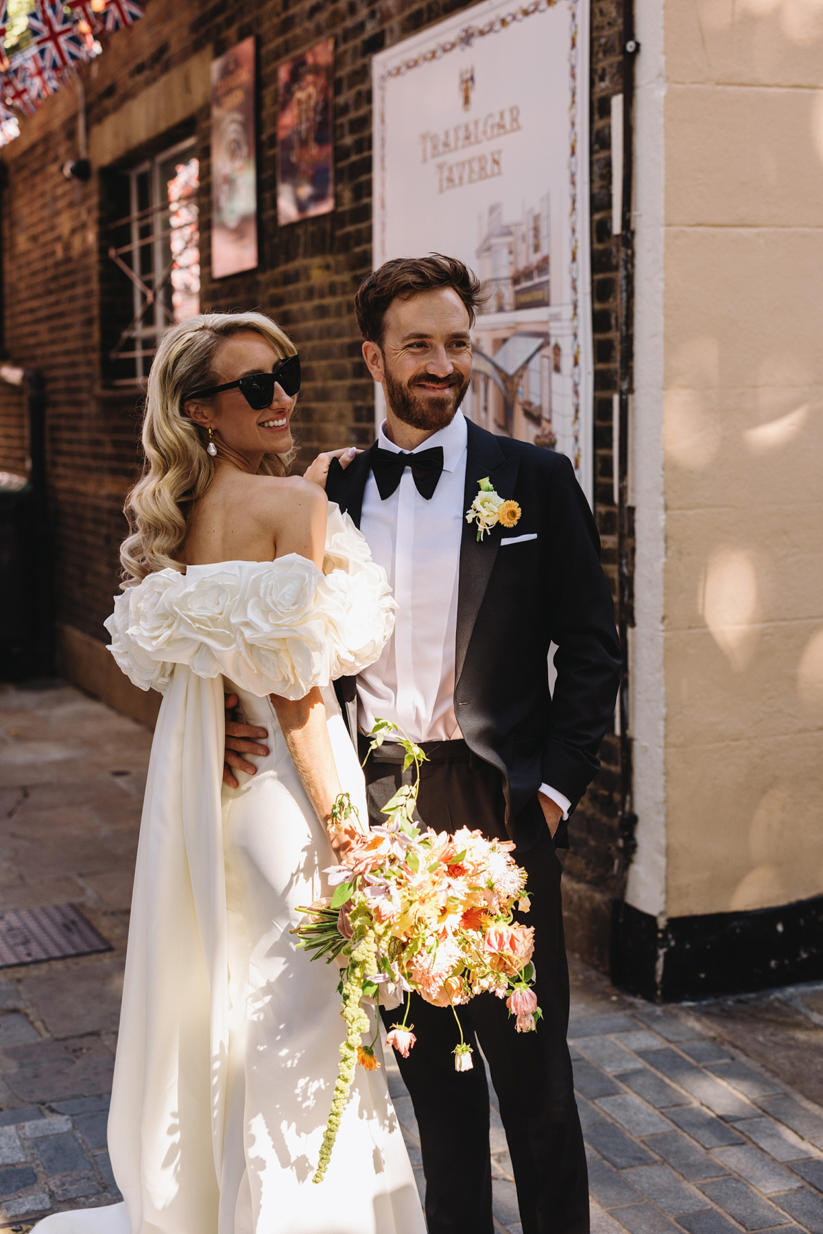 We Invite You To Join The Love My Dress Wedding Directory 1 Bride with long blonde wavy hair wearing an Eva Lendel wedding dress with frilly sleeves. She wears sunglasses and is smiling. Groom in black tie. Wolf & Co Photography.