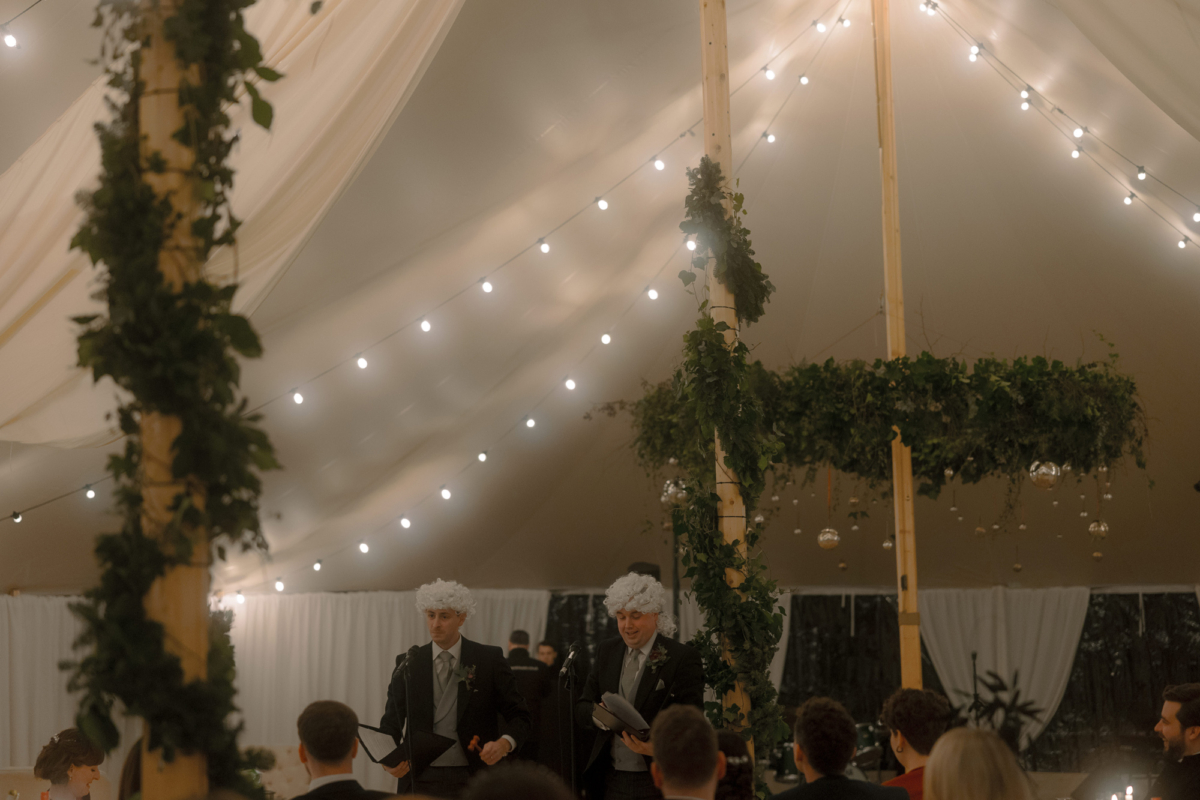 A Movie & Literary Inspired Romantic Christmas Wedding 64 Wedding guests delivering a speech with wigs on, inside a wedding marquee decorated with festoon lights, draping and greenery.