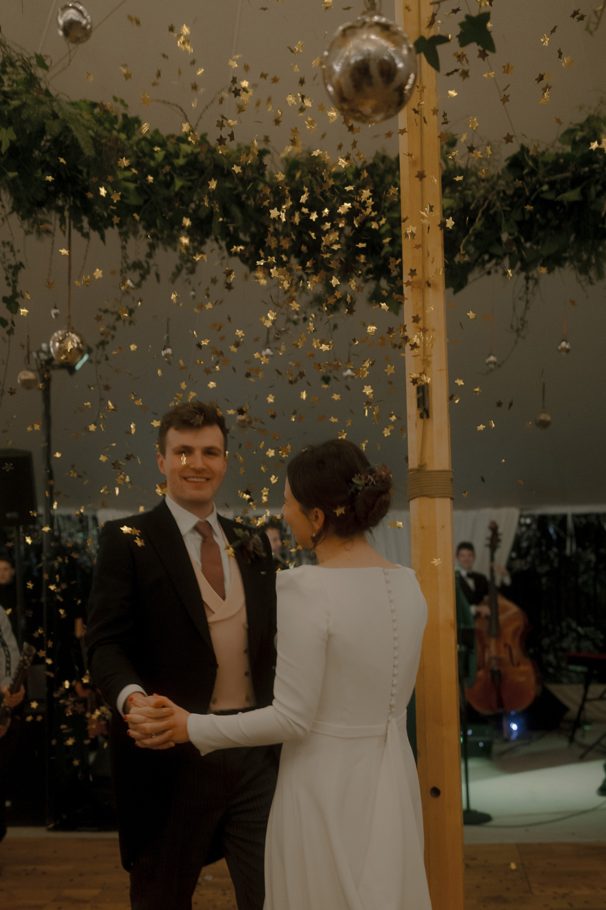 A Movie & Literary Inspired Romantic Christmas Wedding 92 Bride and groom dancing in a wedding marquee whilst being showered with gold star confetti. The bride wears a Suzanne Neville wedding drss.