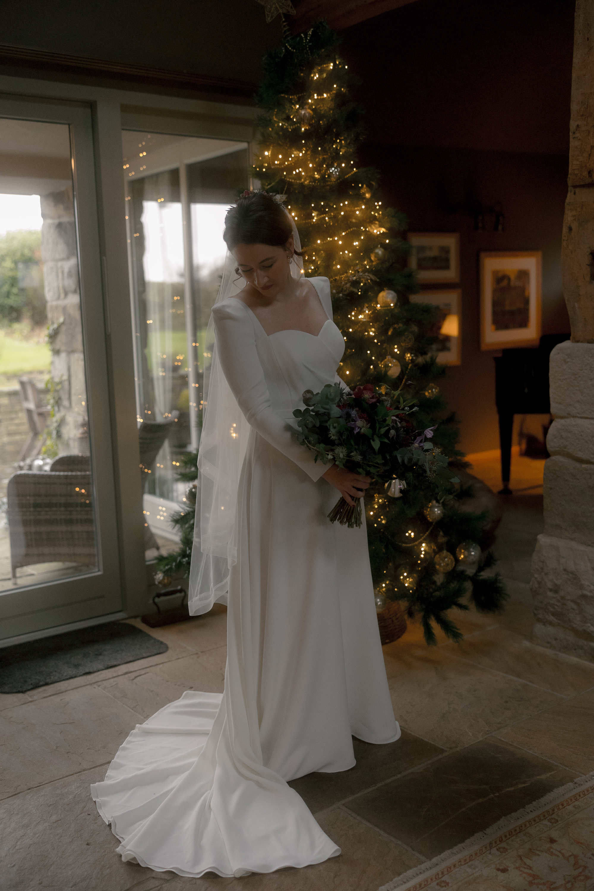 A Movie & Literary Inspired Romantic Christmas Wedding 45 Bride getting ready in the Verdi dress, by Suzanne Neville. She stands next to a Christmas tree and carries a winter wedding bouquet.