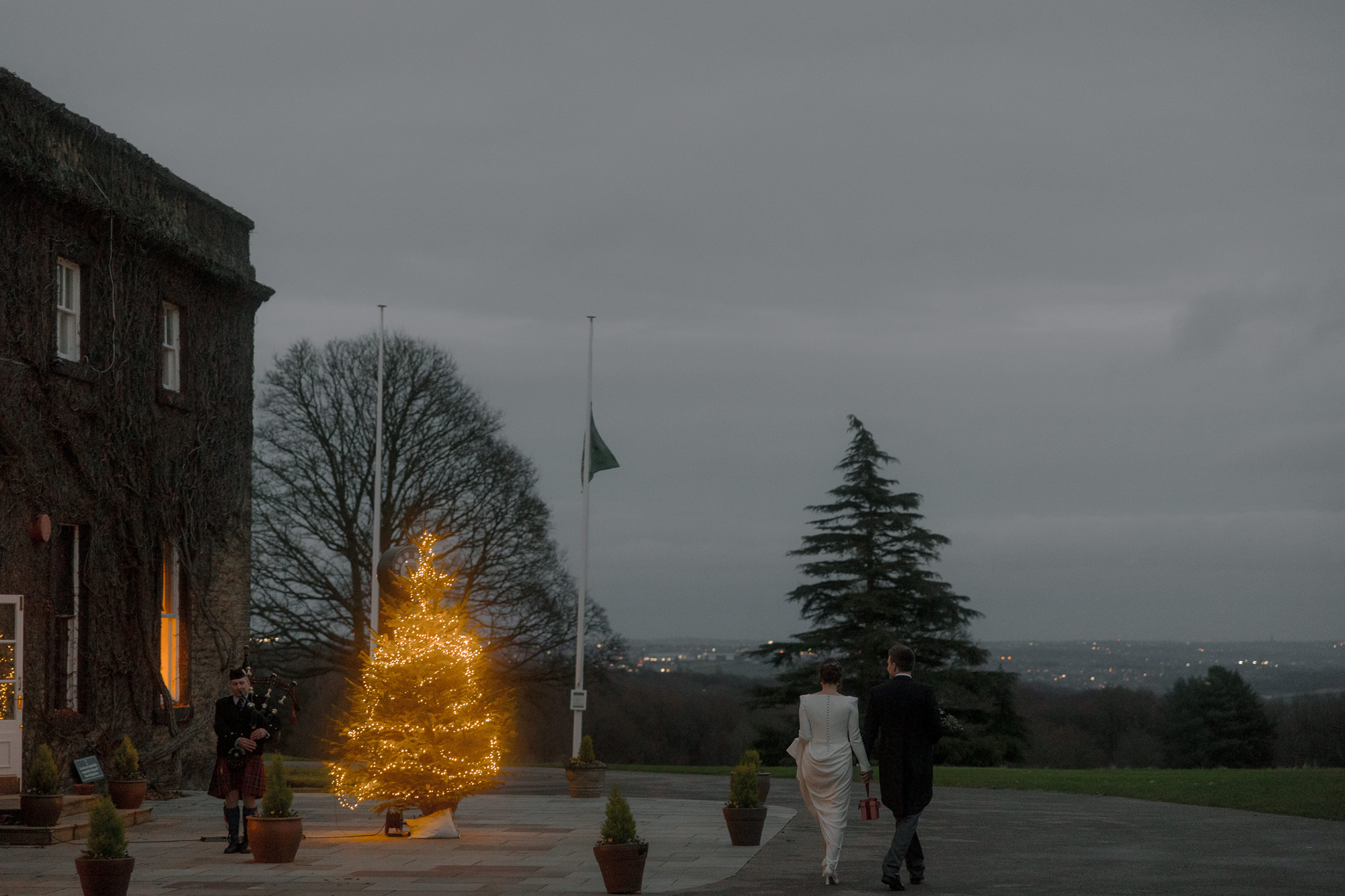 A Movie & Literary Inspired Romantic Christmas Wedding 57 Bride and groom walking towards Fixby Hall with a tree lit with fairylights by the entrance on their winter Christmas wedding day.