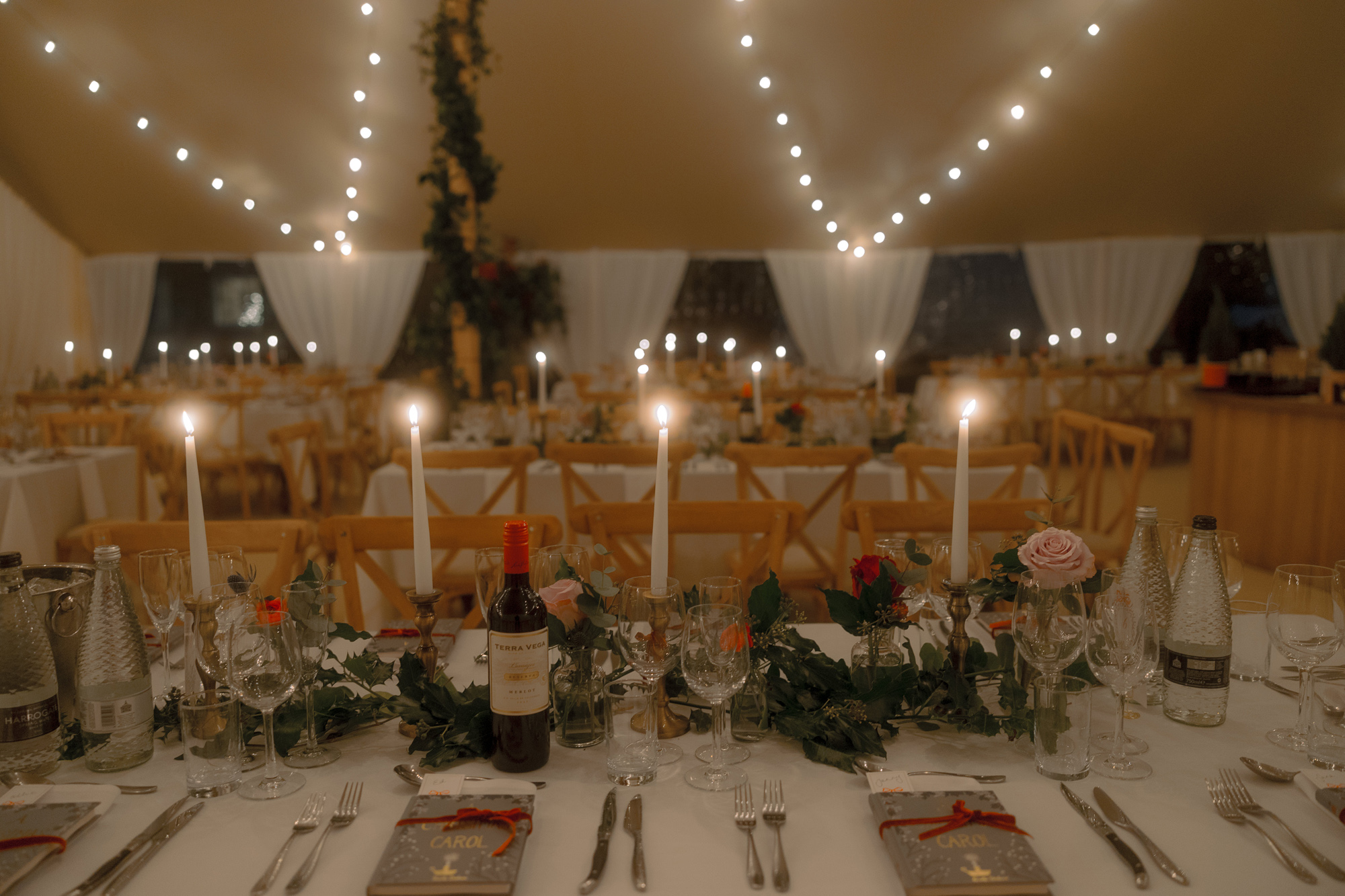 A Movie & Literary Inspired Romantic Christmas Wedding 63 Christmas wedding table decor inside a marquee. With taper candles, festoon lights, red and pink roses and guests gifted with a copy of 'A Christmas Carol' book.