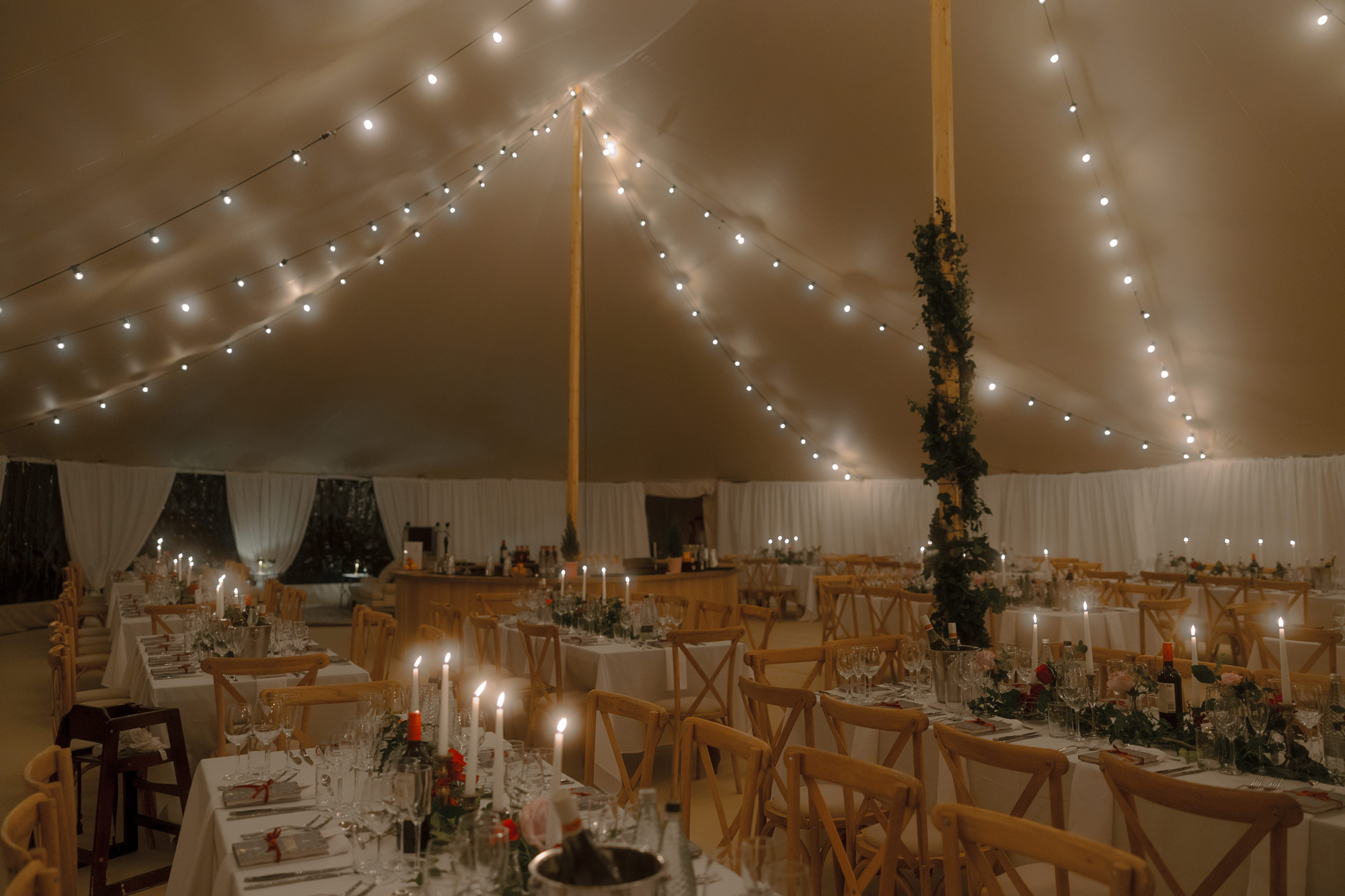 A Movie & Literary Inspired Romantic Christmas Wedding 58 Wedding marquee lit in warm light of taper candles and festoon lights. Poles are decorated with greenery. Cross back chairs and long trestle tables.