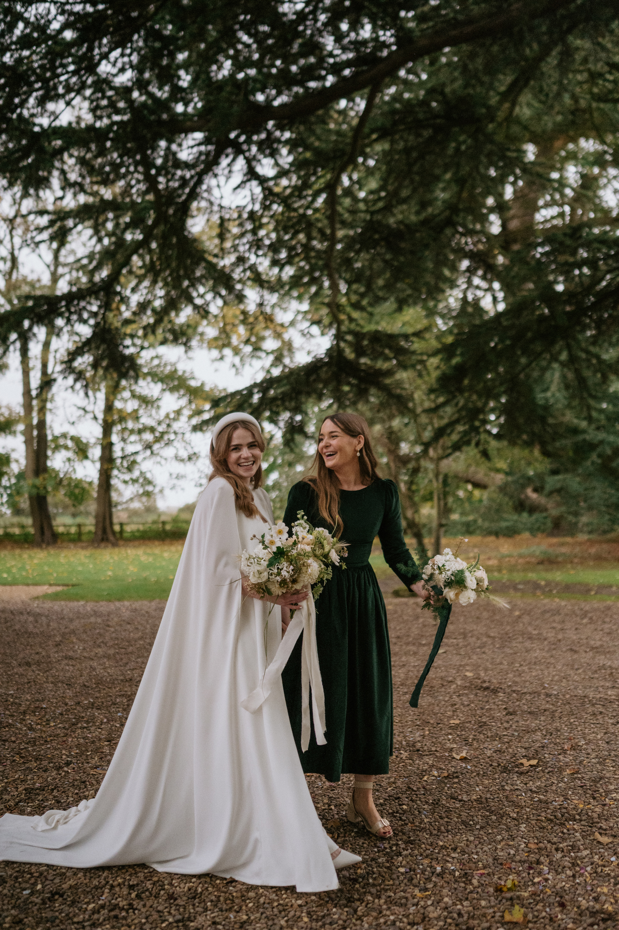 Bride wearing a Jesus Peiro wedding dress and cape, carrying an elegant neutral bouquet with silk ribbons, she is laughing with her bridesmaid in a dark green velvet midi dress.
