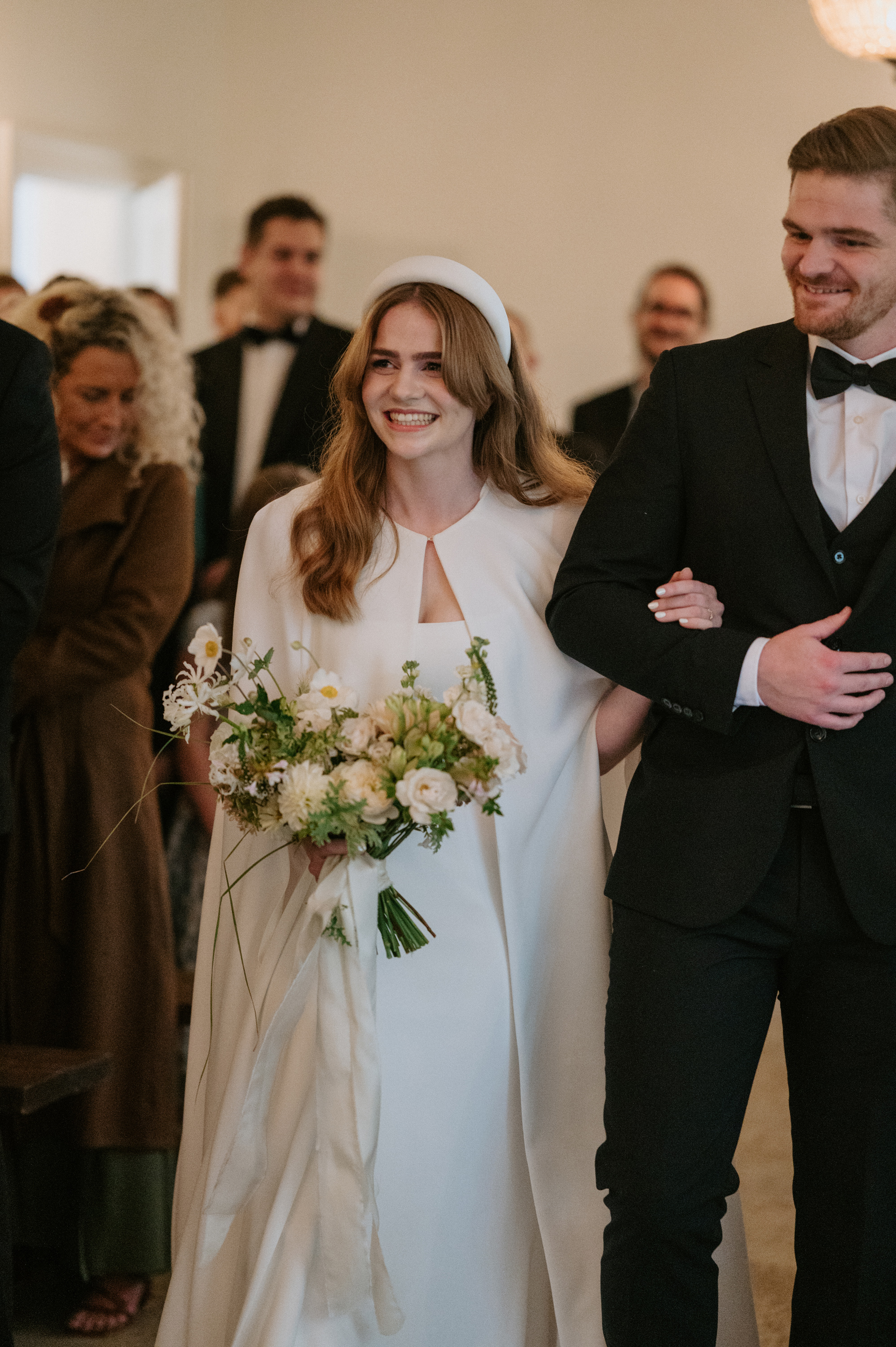 Bride being accompanied by her brother down the aisle. She wears a Jesus Peiro wedding dress and cape and carries an elegant neutral bouquet with silk ribbons. 