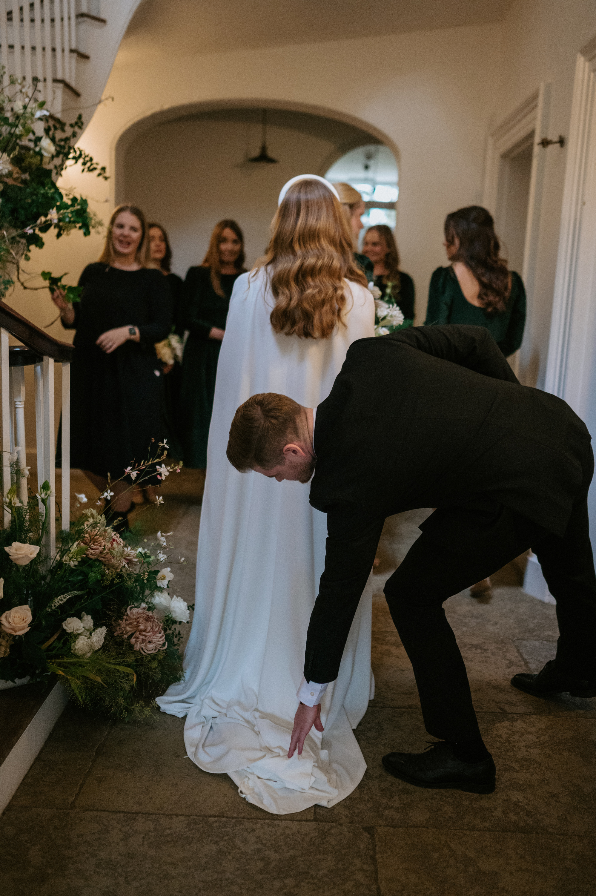 Phoebe in Jesus Peiro for her Elegant November Wedding at Aswarby Rectory 22 Bride in a Jesus Peiro cape and headband having her dress train adjusted by her brother as her bridesmaids greet her at the foot of the stairs.