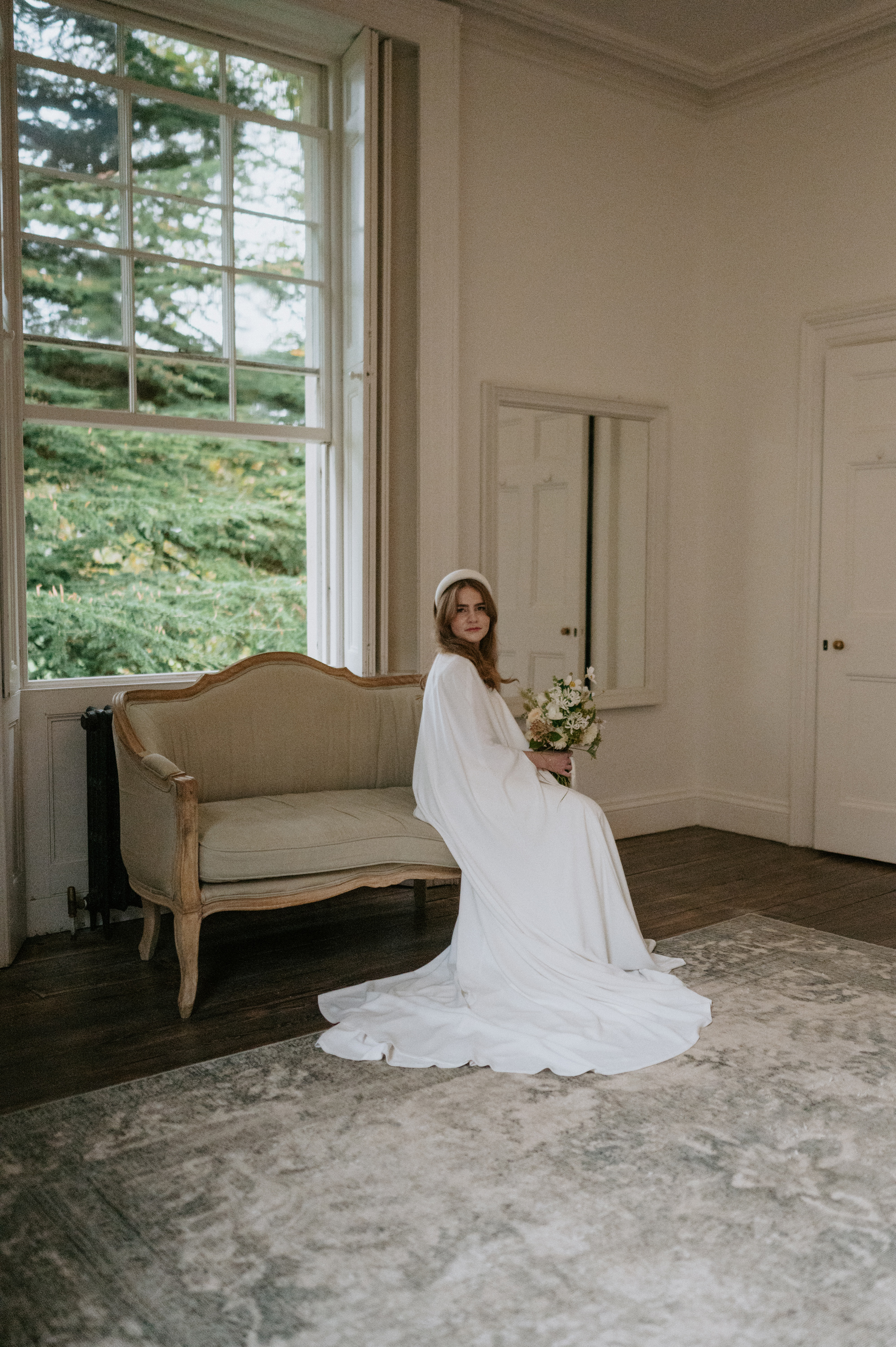 Phoebe in Jesus Peiro for her Elegant November Wedding at Aswarby Rectory 21 Bride in a Jesus Peiro cape and headband sat on a chic vintage sofa by a large window, inside Aswarby Rectory.