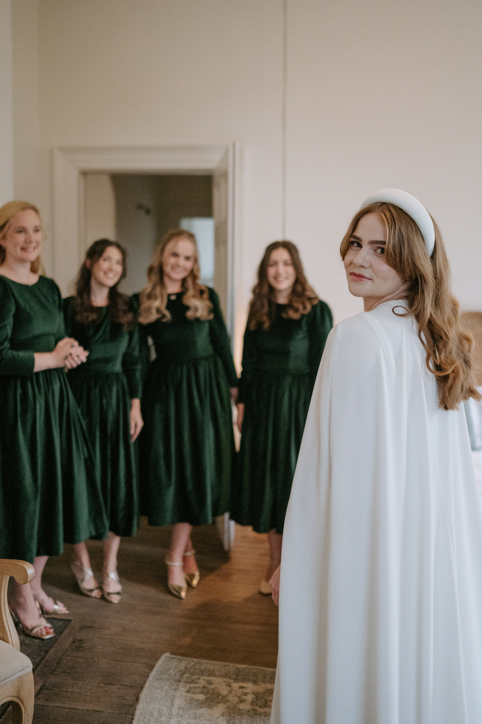 Bride wearing a Jesus Peiro headband and cape. Bridesmaids in the background wearing dark green velvet dresses.