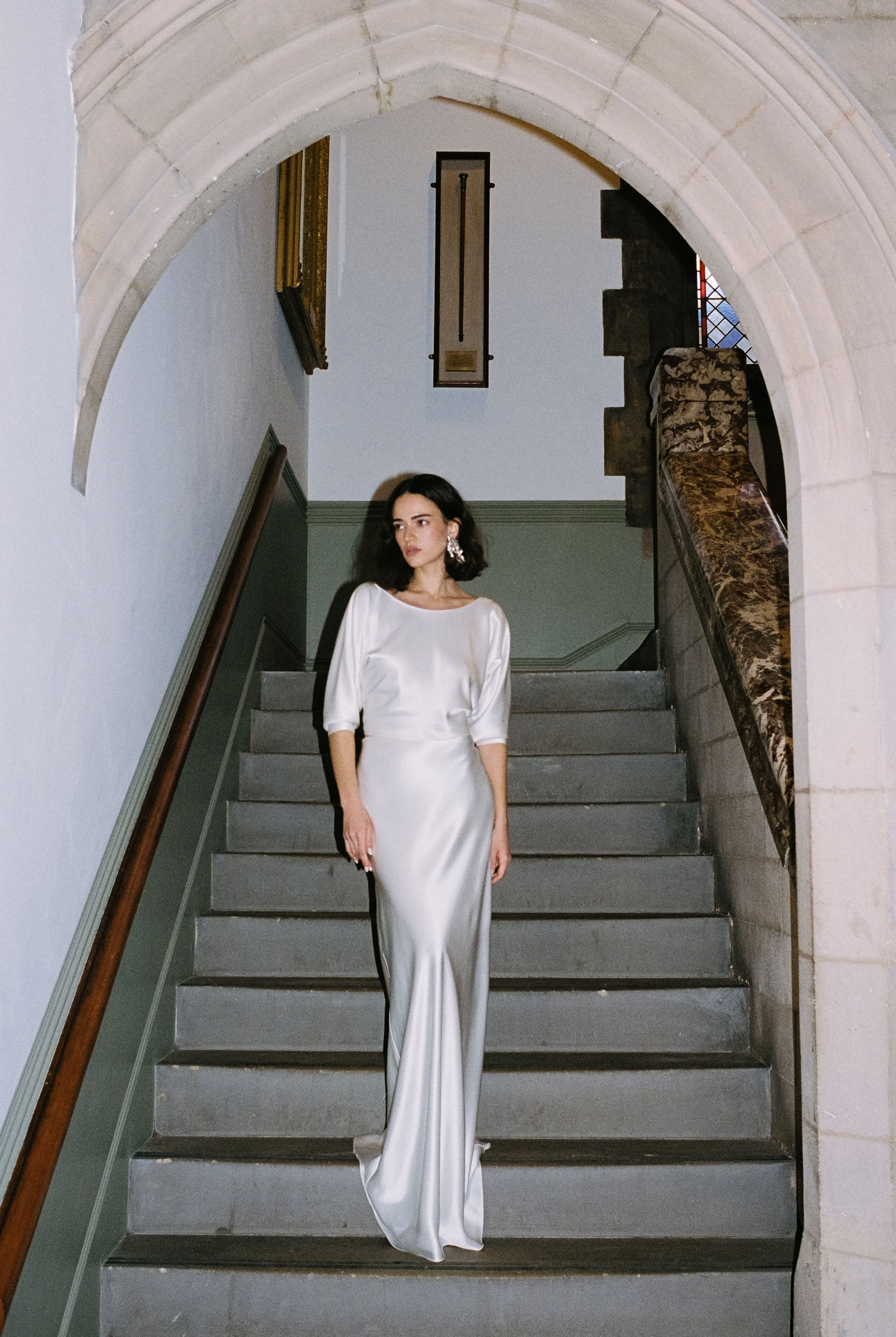 The Juliet Collection, by Rolling In Roses 16 Elegant bride standing on a flight of stairs wearing a silk bias cut wedding dress with elbow length sleeves by Rolling in Roses.