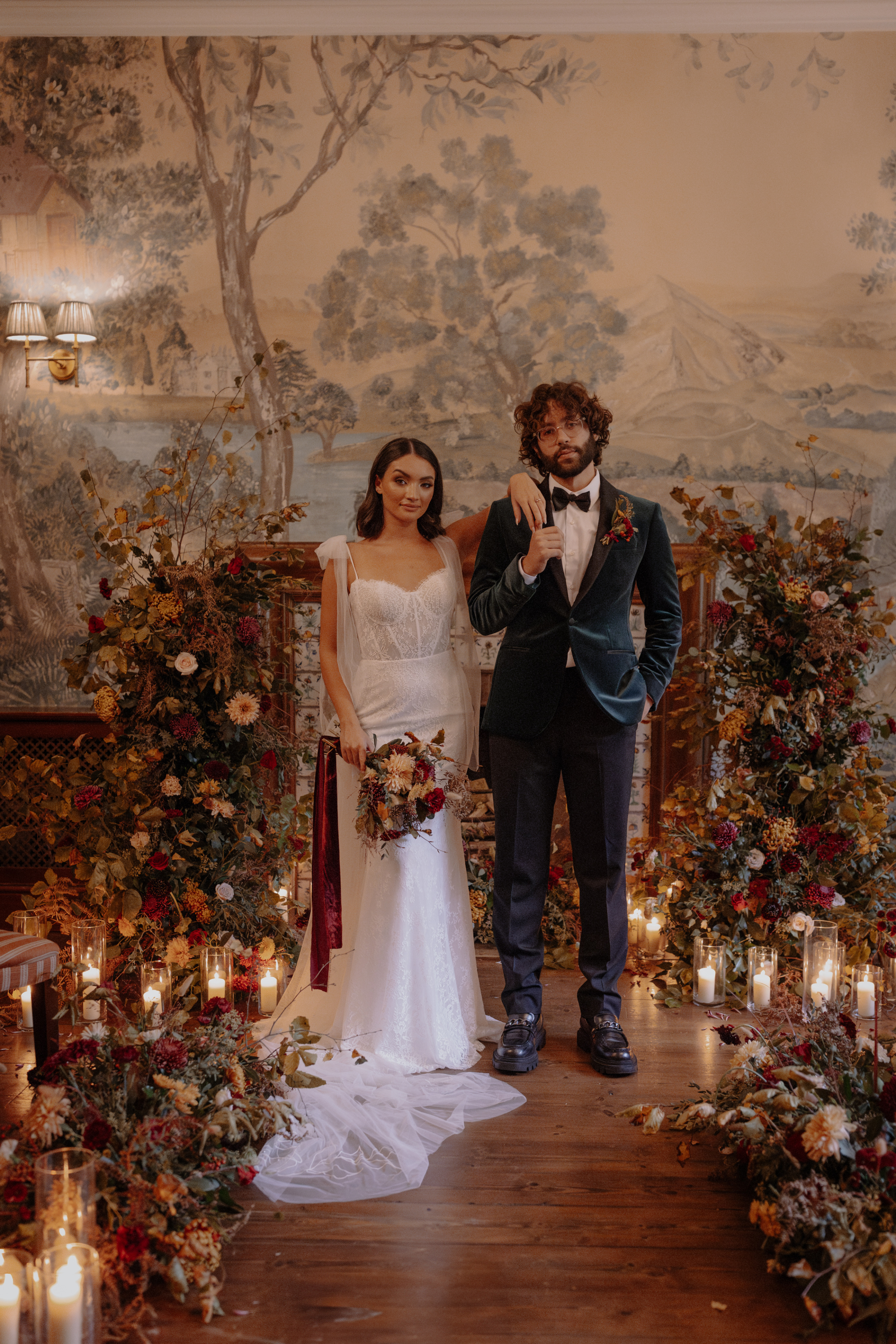 A Little Wild, A Little Luxe: Helen Rose Photography's Beautiful Peak District Editorial 6 Bride standing with her arm raised and her hand rested on her groom's shoulder - he holds her fingers as they hang down his lapel. He wears a blue velvet suit. They stand by a fireplace that has been surrounded by Autumnal floral styling and an abundance of candles.