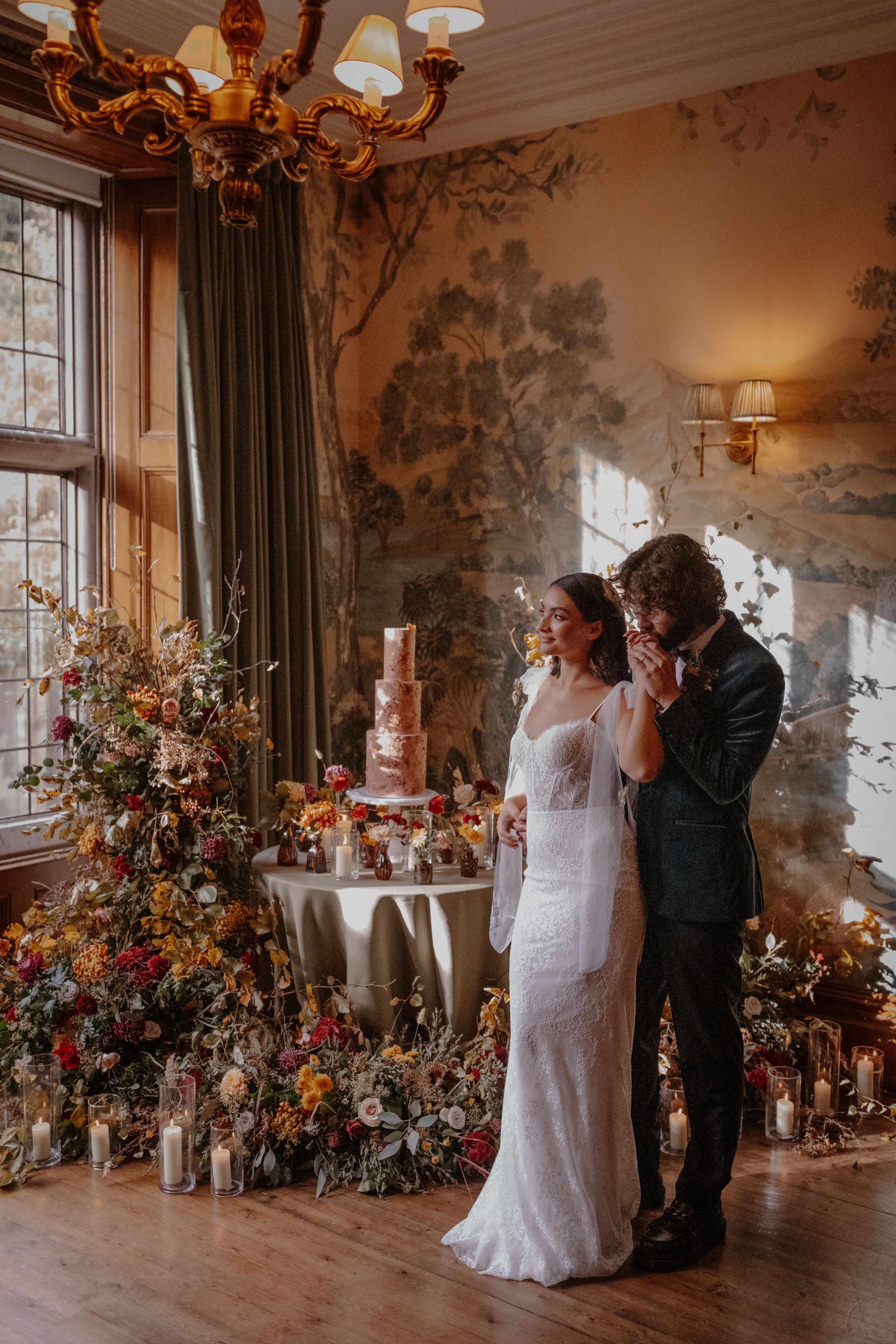 Bride and groom standing in a grand room next to a small round table with a tall 3 tier modern wedding cake that is surrounded by an abundance of Autumn wedding flowers and candles.