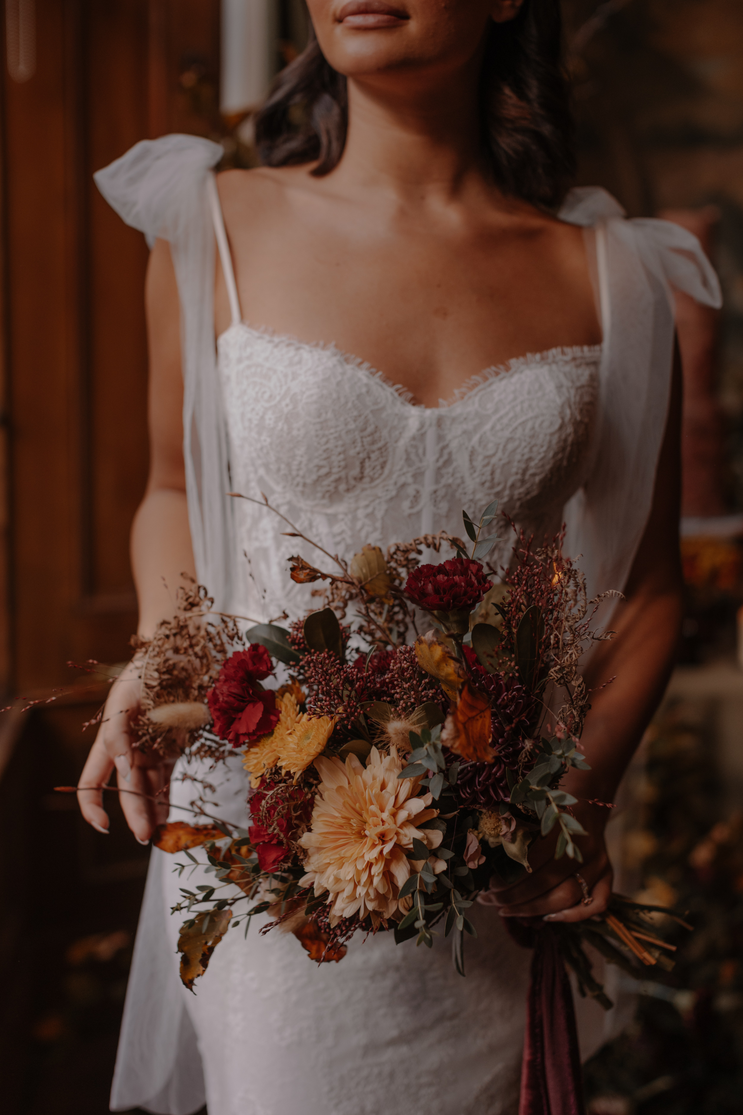 Close up shot of a bride clutching an Augumn wedding bouquet. She wears a dress with shoulder ribbons.