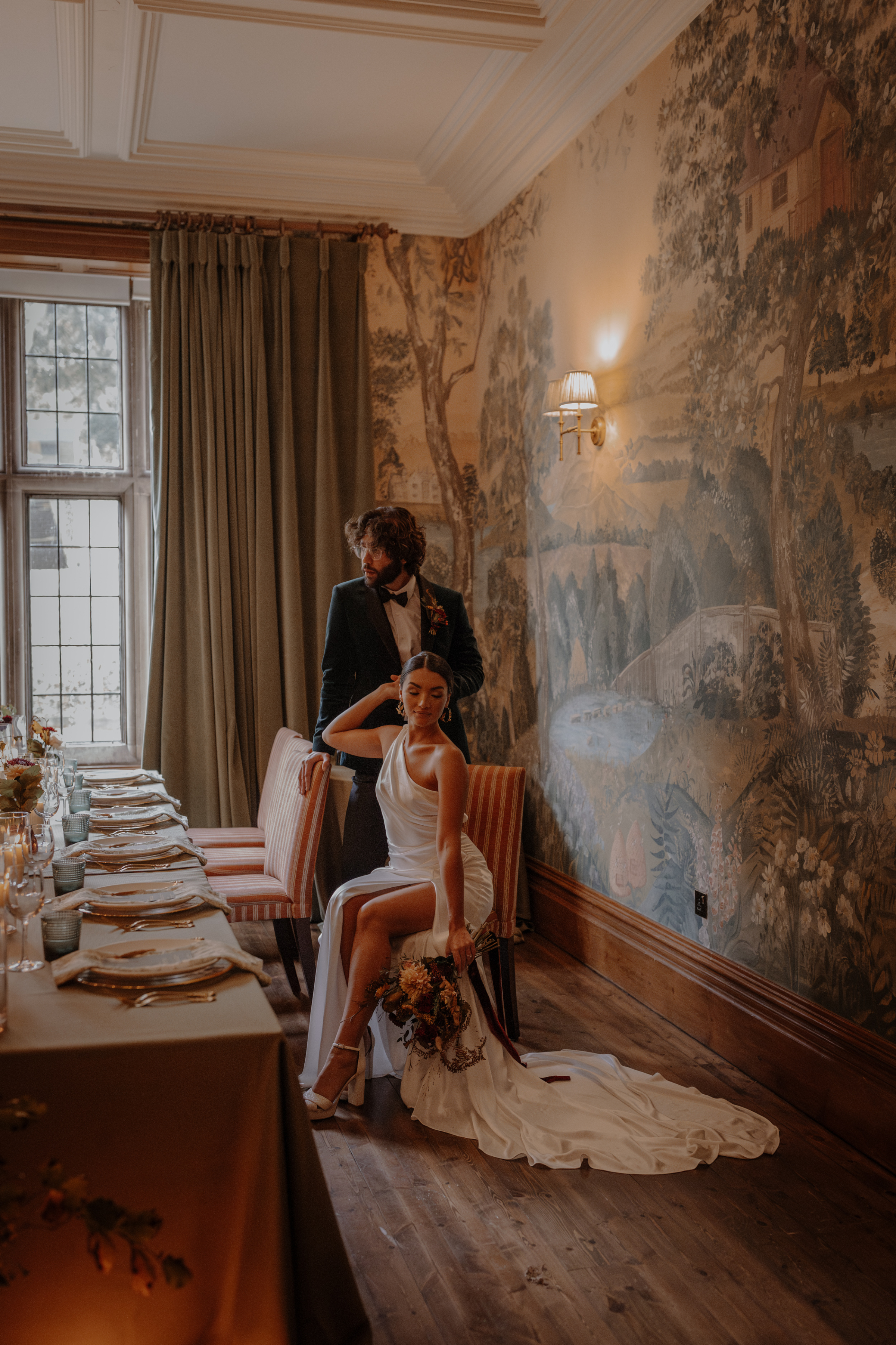 Bride and groom sat in a grand room by a wedding table - the bride wears a sensuous wedding dress that reveals her thigh - she is sat down and the dress rain is elegantly puddled  around her chair.