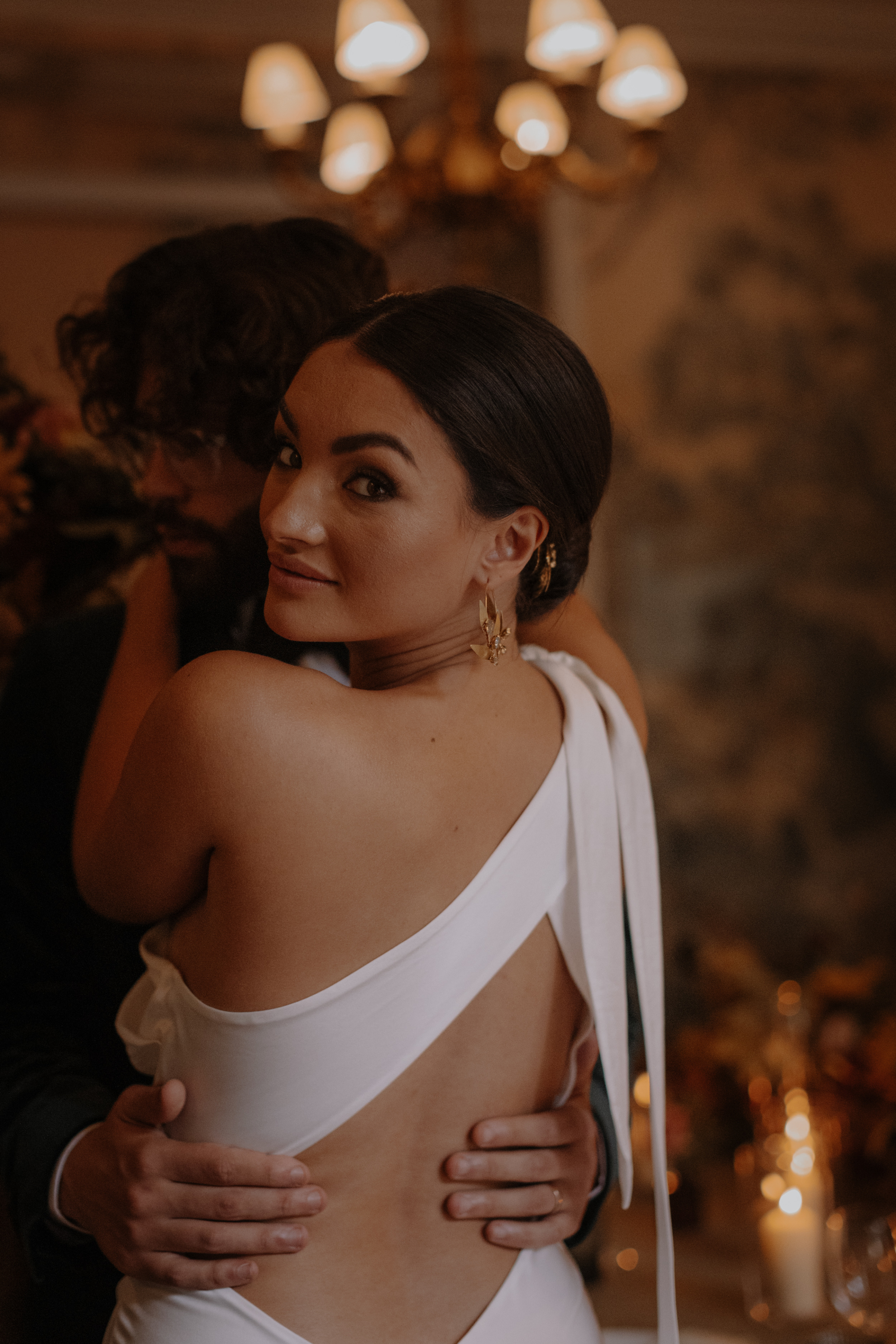 Bride wearing a sensuous modern wedding dress  with a strap that crosses her back and ties on one shoulder. She wears modern gold jewellery and has her hair up on low bun.