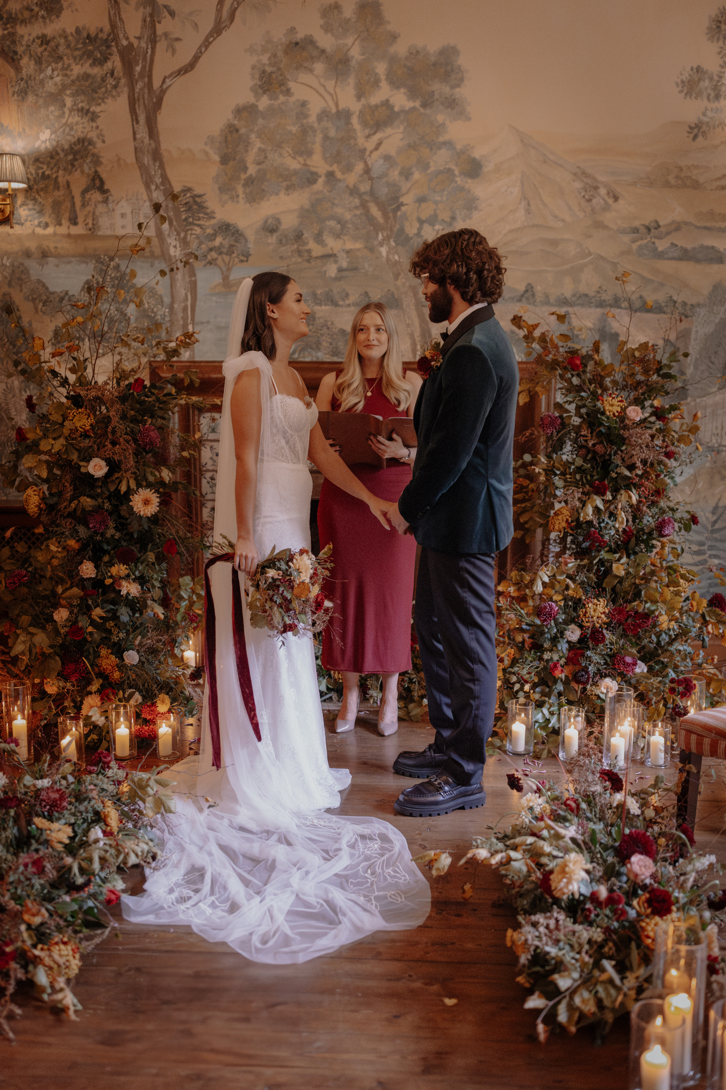 A Little Wild, A Little Luxe: Helen Rose Photography's Beautiful Peak District Editorial 17 Bride and groom during their wedding ceremony by a fire that is surrounded by Autumn flowers and a broken floral arch. There is an abundance of blooms and candles all around. A celebrant in a maroon dress leads the ceremony.