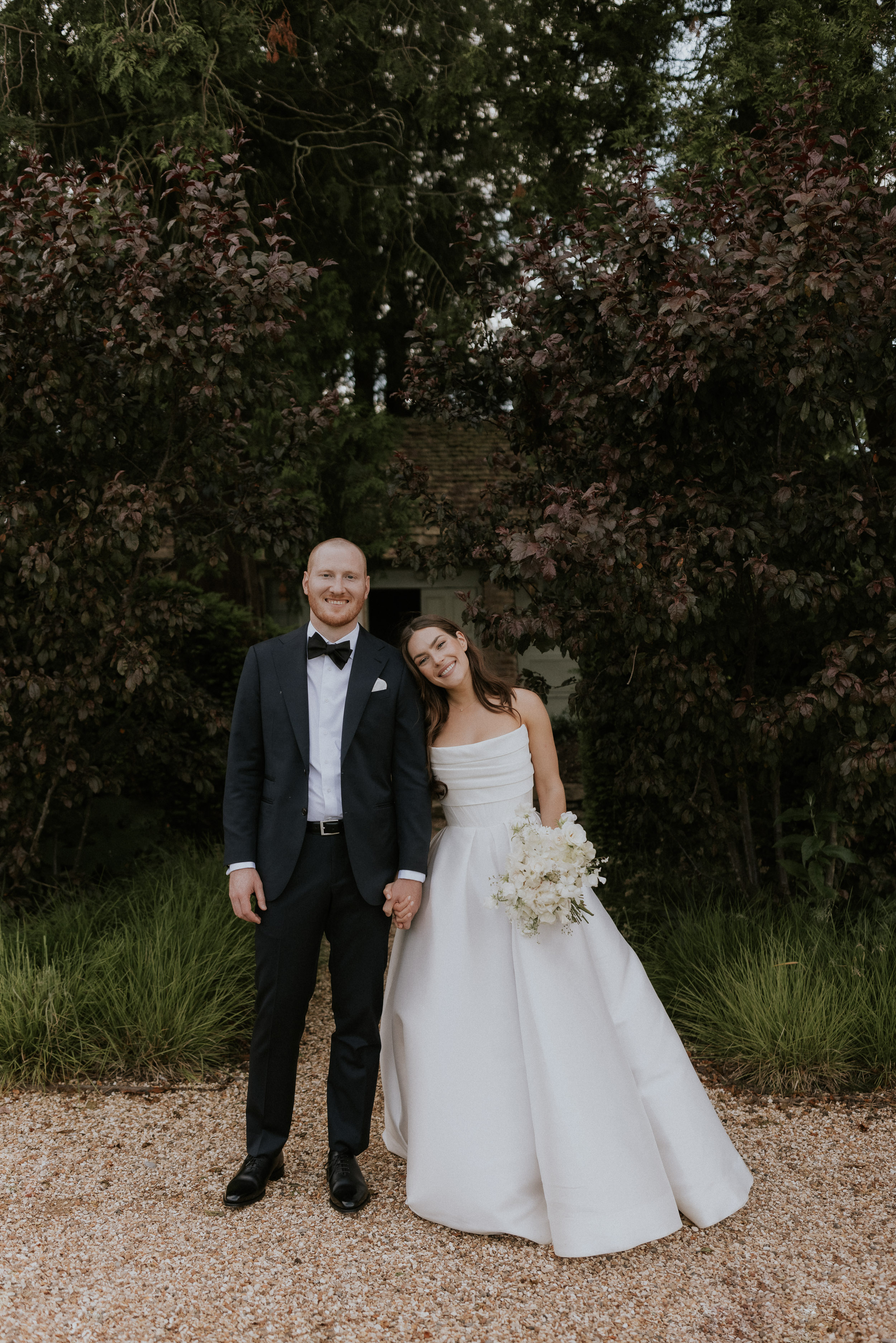 A Romantic Summer Wedding at Kin House in Wiltshire 38 Bride wearing Alex Perry and clutching a medium size bouquet of all white flowers. She ls leaning in to her groom who wears black tie tuxedo. Maja Tsolo Photography.