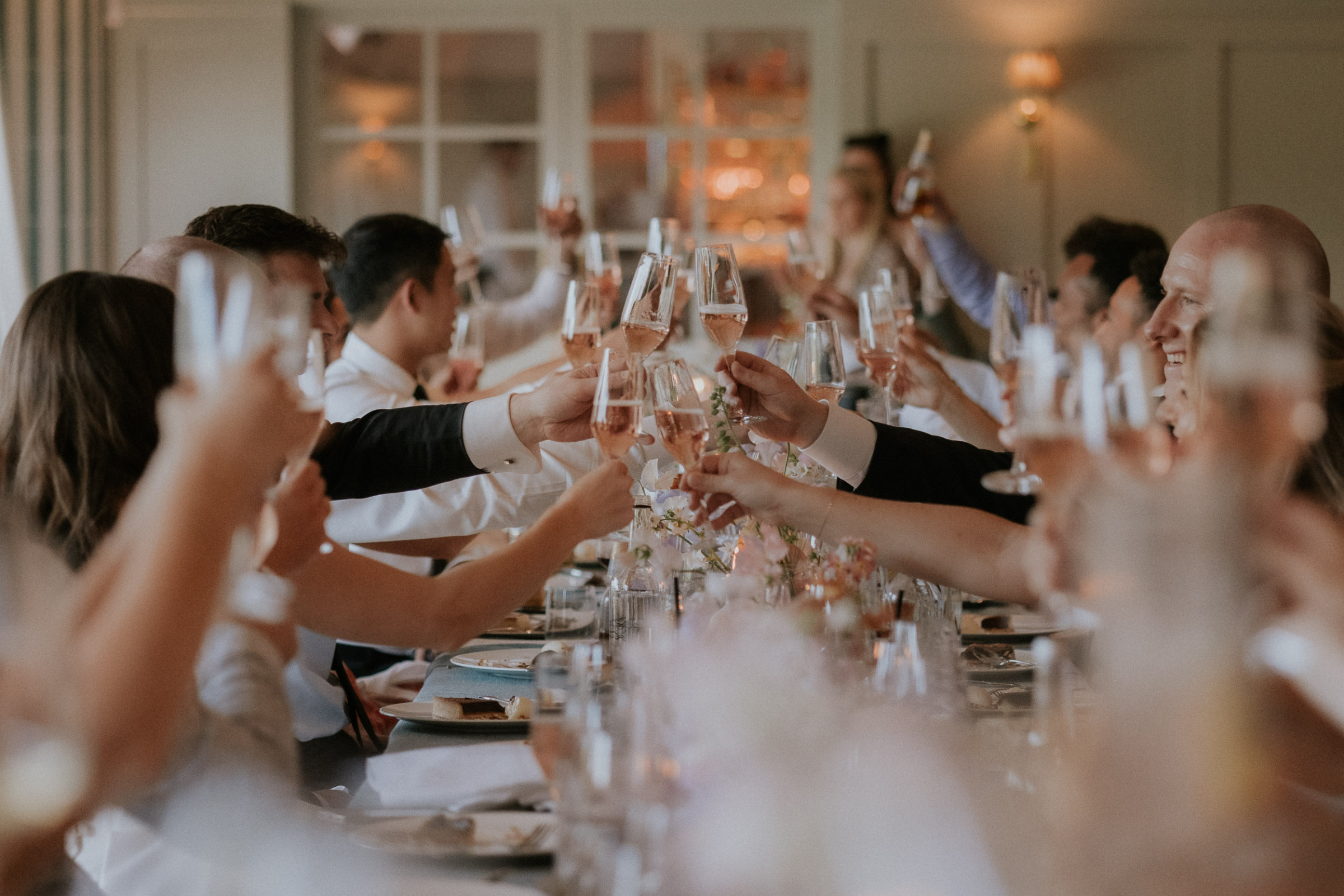 A Romantic Summer Wedding at Kin House in Wiltshire 53 Guests cheersing their glasses over the table at a wedding. Maja Tsolo Photography.