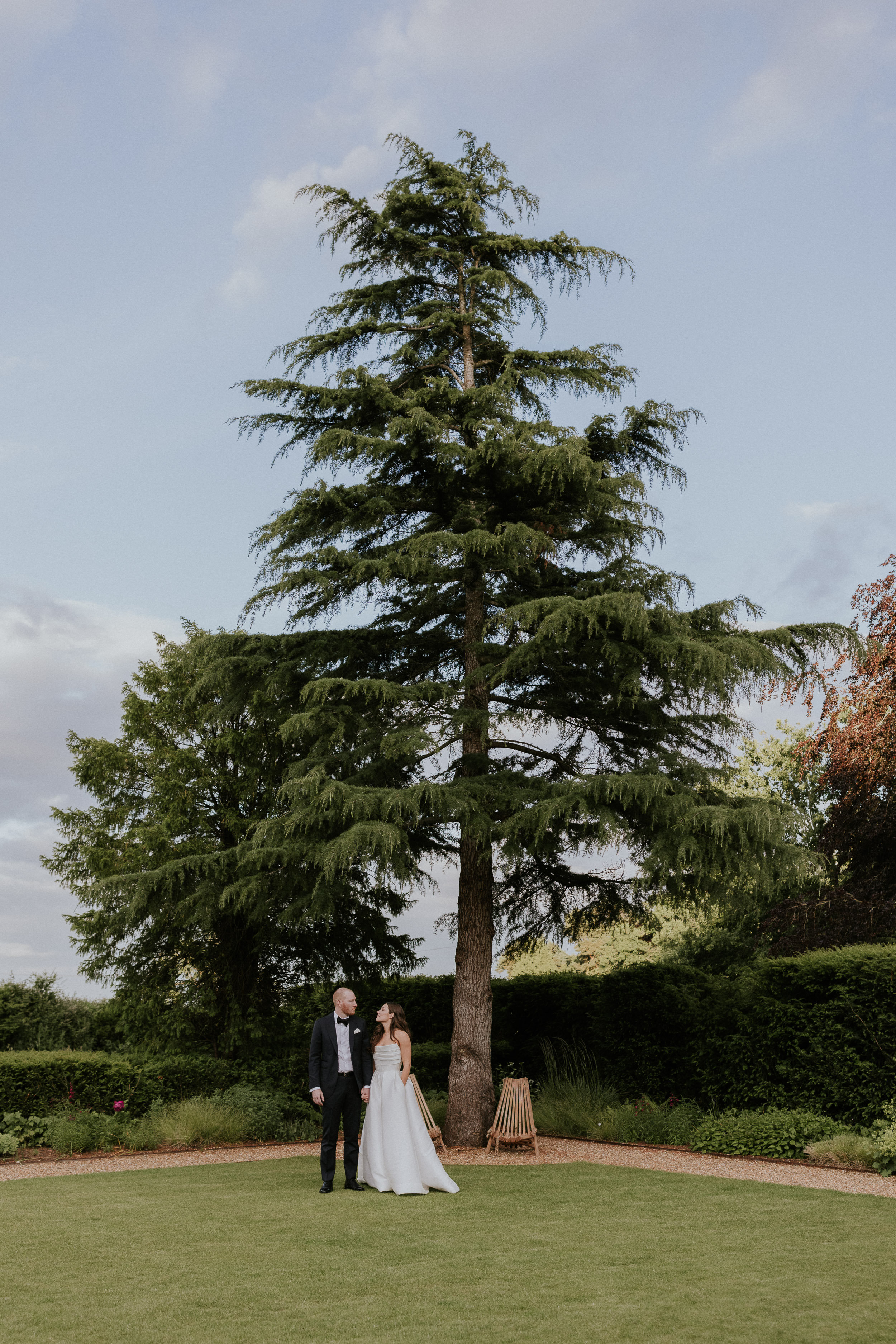 Bride and groom photographed at a distance with a tall conifer in the background at Kin House, Wiltshire. Maja Tsolo Photography.