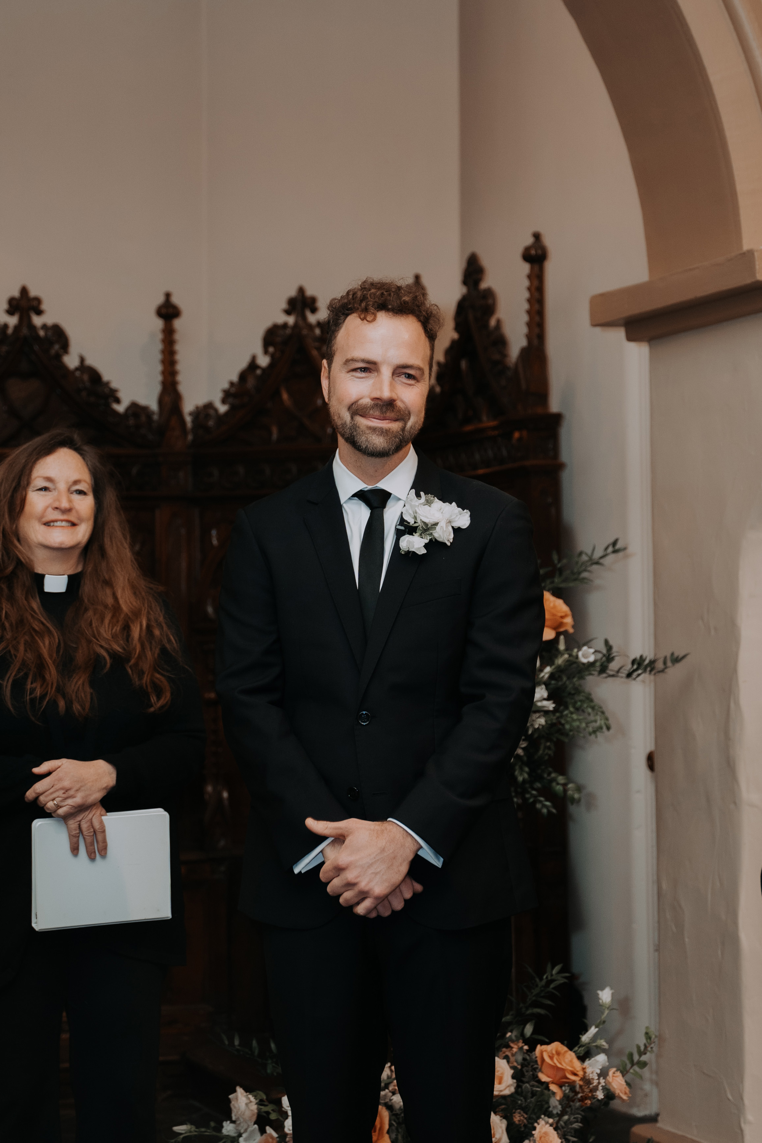 Groom smiling towards his wife as she walks down the aisle. Behind him stands a female vicar smiling also.