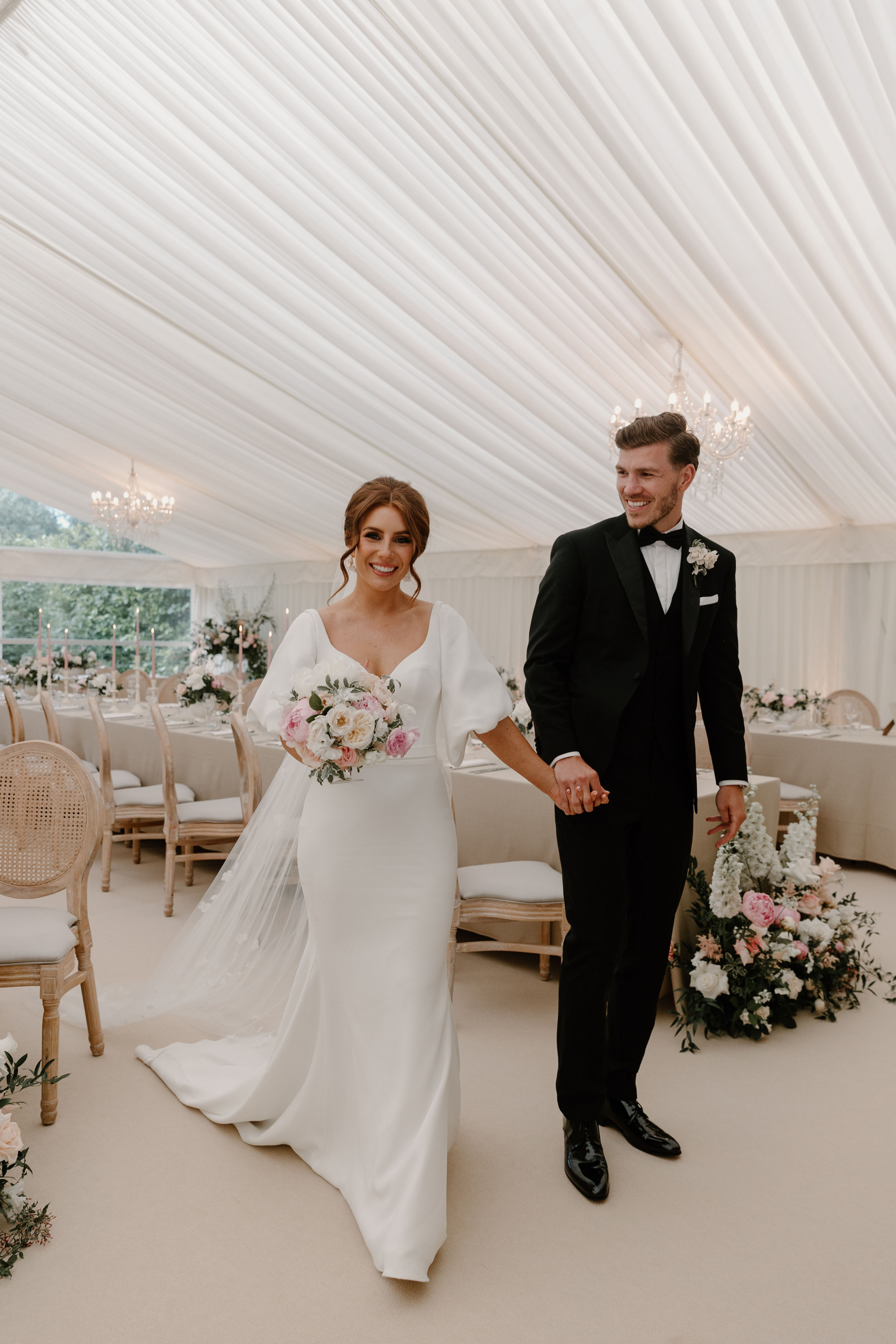 Groom in black tie and bride in Suzanne Neville wedding dress walking through their wedding marquee.