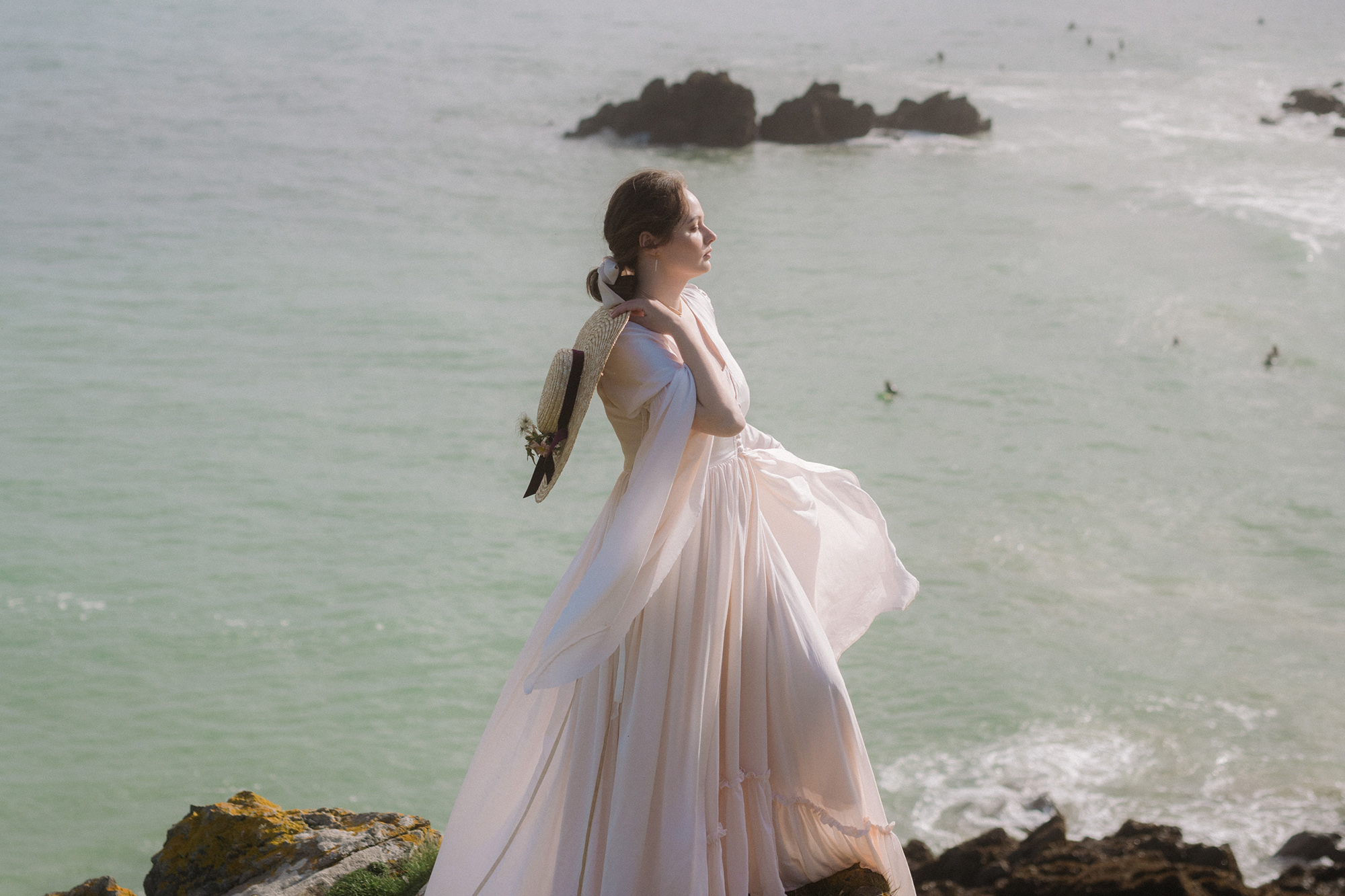 Bride standing on a cliff in Cornwall in the breeze holding a hat across her shoulder and wearing a ruffled dress that is floating in the breeze.