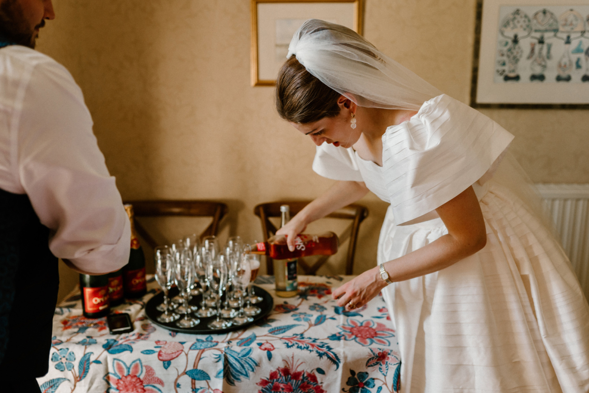 Hannah in Jesus Peiro for her Intimate Brunton House Wedding in Northumberland 38 Bride wearing a puff sleeve Jesus Peiro wedding dress and veil over her bun, is leaning over to pour fizz into tall stemmed glasses on her wedding day.