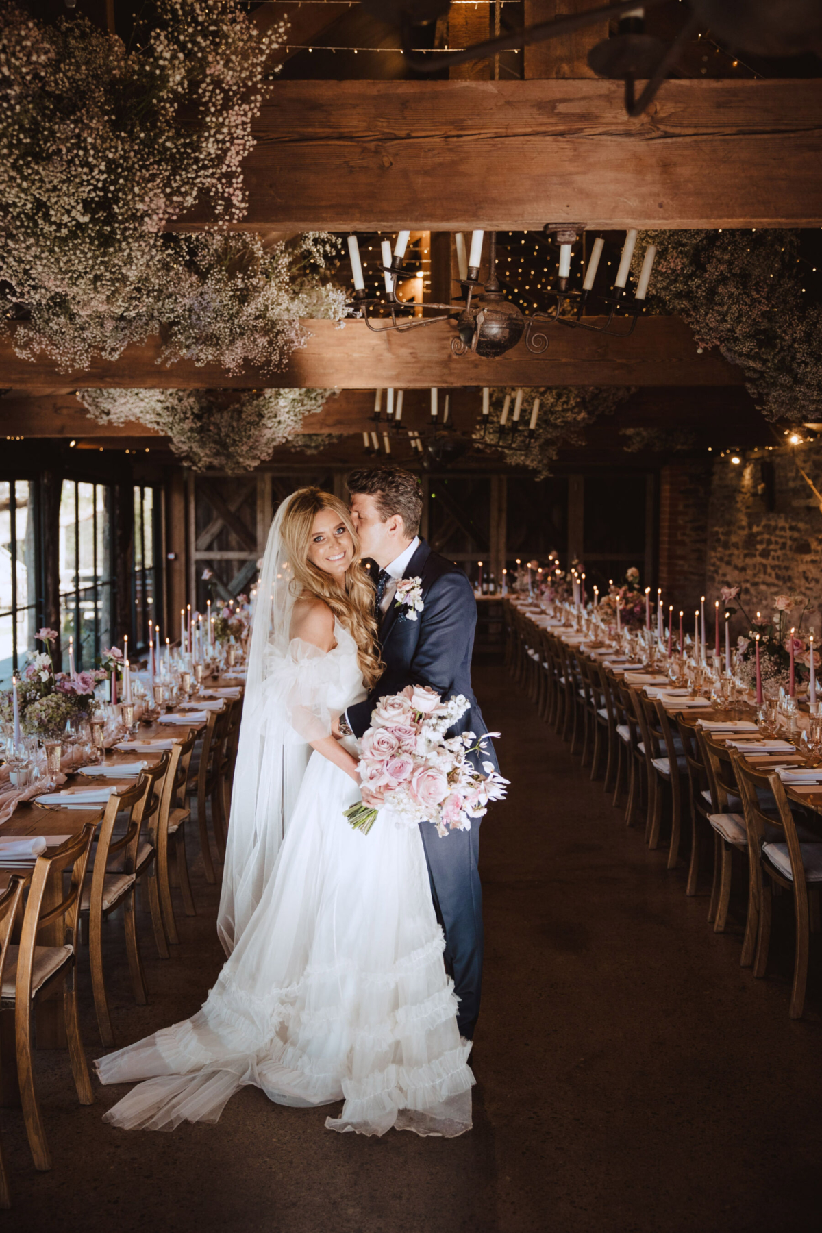 Discover the Magic of Dewsall Court at Their Exclusive, By Appointment Only, Open House, 26th-27th April 4 Dewsall Court, Herefordshire wedding venue. Bride and groom in a barn with gypsophila hanging from the beams and tall pink and red taper candles on long trestle tables.