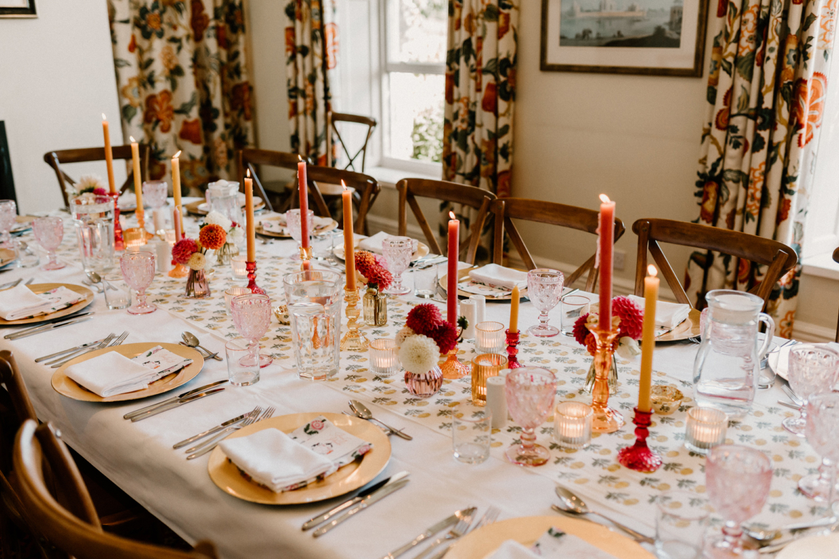 Hannah in Jesus Peiro for her Intimate Brunton House Wedding in Northumberland 39 Wedding reception table setting featuring patterned floral table runner, peach charger plates, floral napkins, and tall taper candles in pale red and peach. Soft pink glasses and cross back chairs.