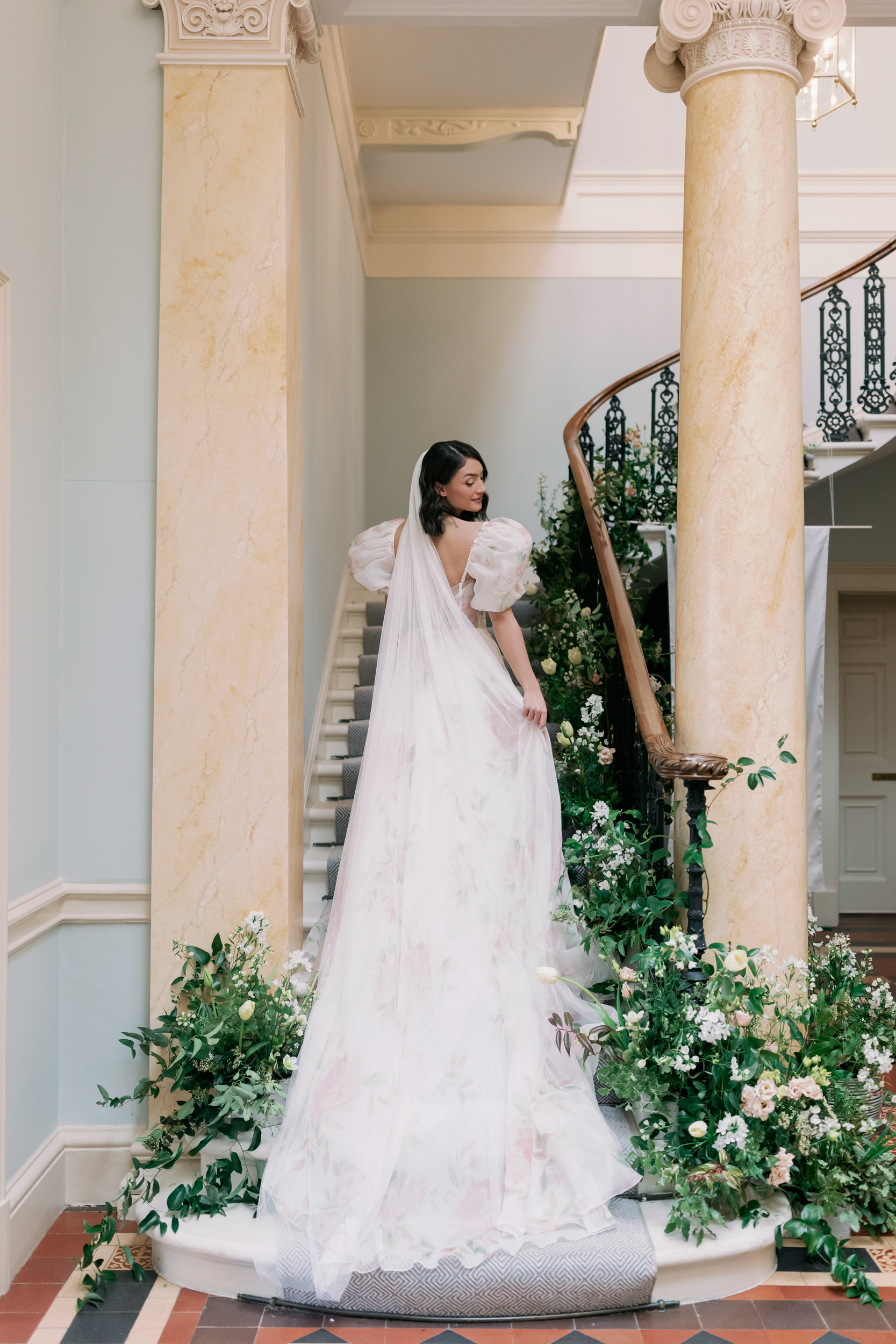More Is More: Wedding Design That is Elegantly & Effortlessly Extra 9 Bride wearing a puff sleeved floral wedding dress and standing at the foot of a sweeping staircase decorated with flowers.