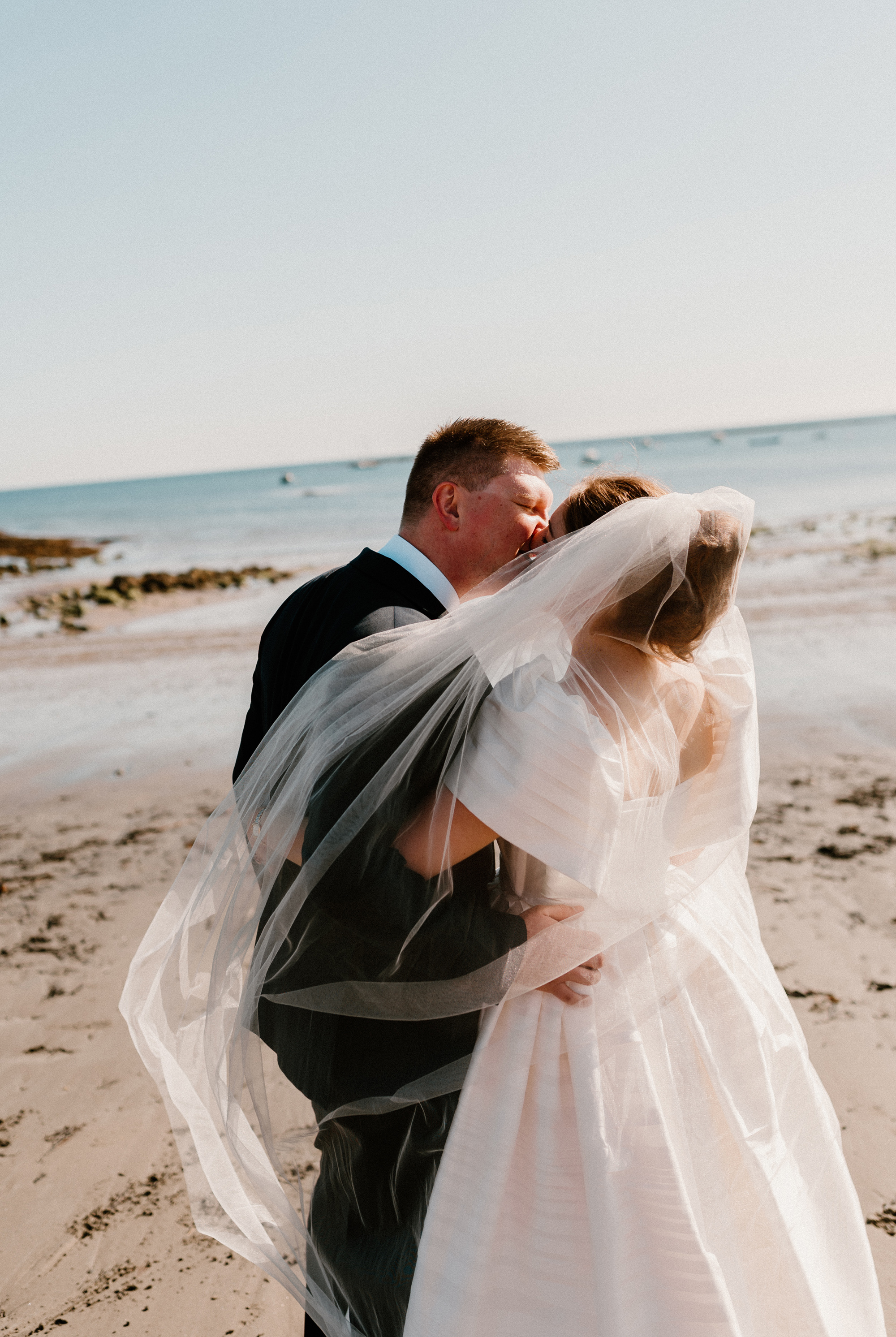 Hannah in Jesus Peiro for her Intimate Brunton House Wedding in Northumberland 47 Bride in a puff sleeve Jesus Peiro wedding dress and veil being swept in the breeze kissing her groom on the beach.