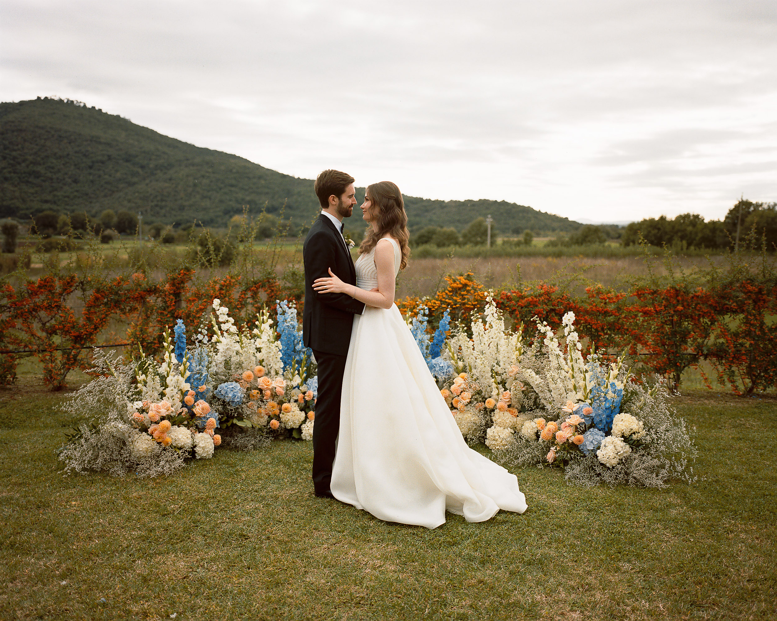 Bride and groom standing by a meadow inspired floral installation outdoors. Relais La Corte Dei Papi Wedding in Cortona, Italy