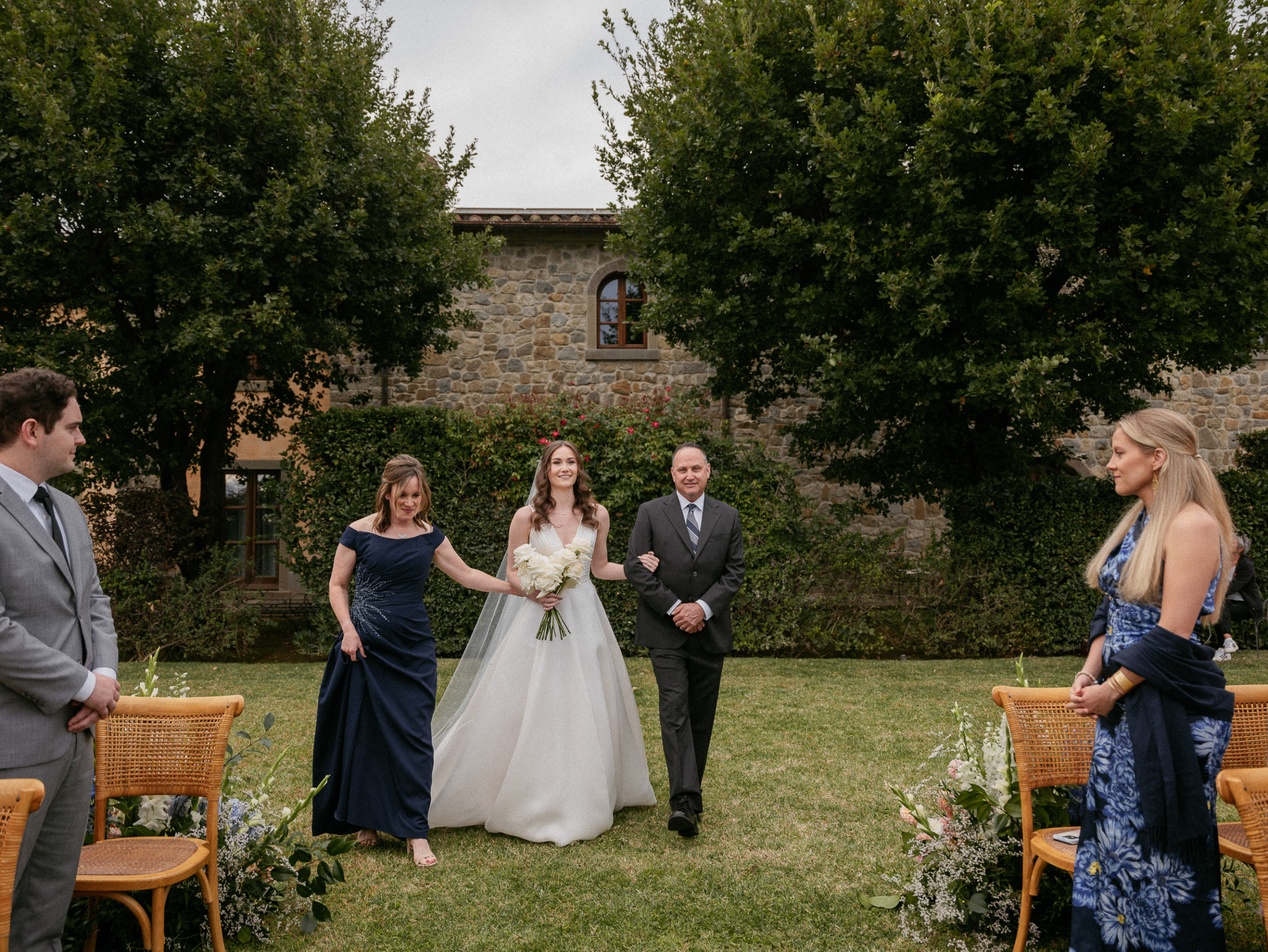 Bride being accompanied by her parents to her  outdoor Italian wedding ceremony at  Relais La Corte Dei Papi in Cortona in Italy.