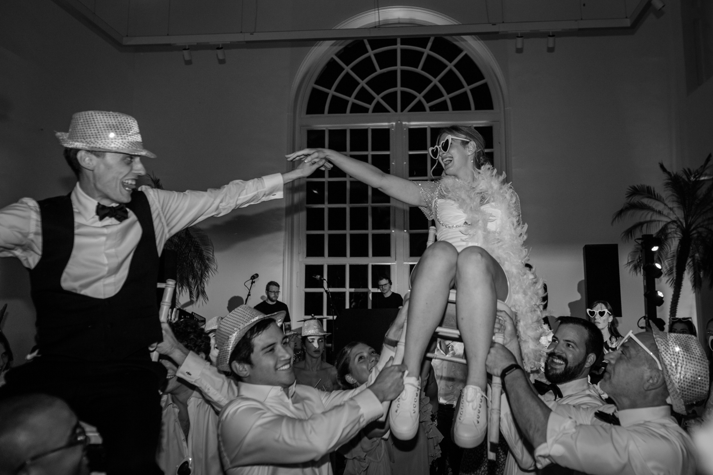 A Dreamy Kew Gardens Glasshouse Wedding Planned by Rachel Dalton 46 Jewish wedding with the bride being lifted on a chair - she wears a feather boah, heart sunglasses, a short wedding dress and trainers.