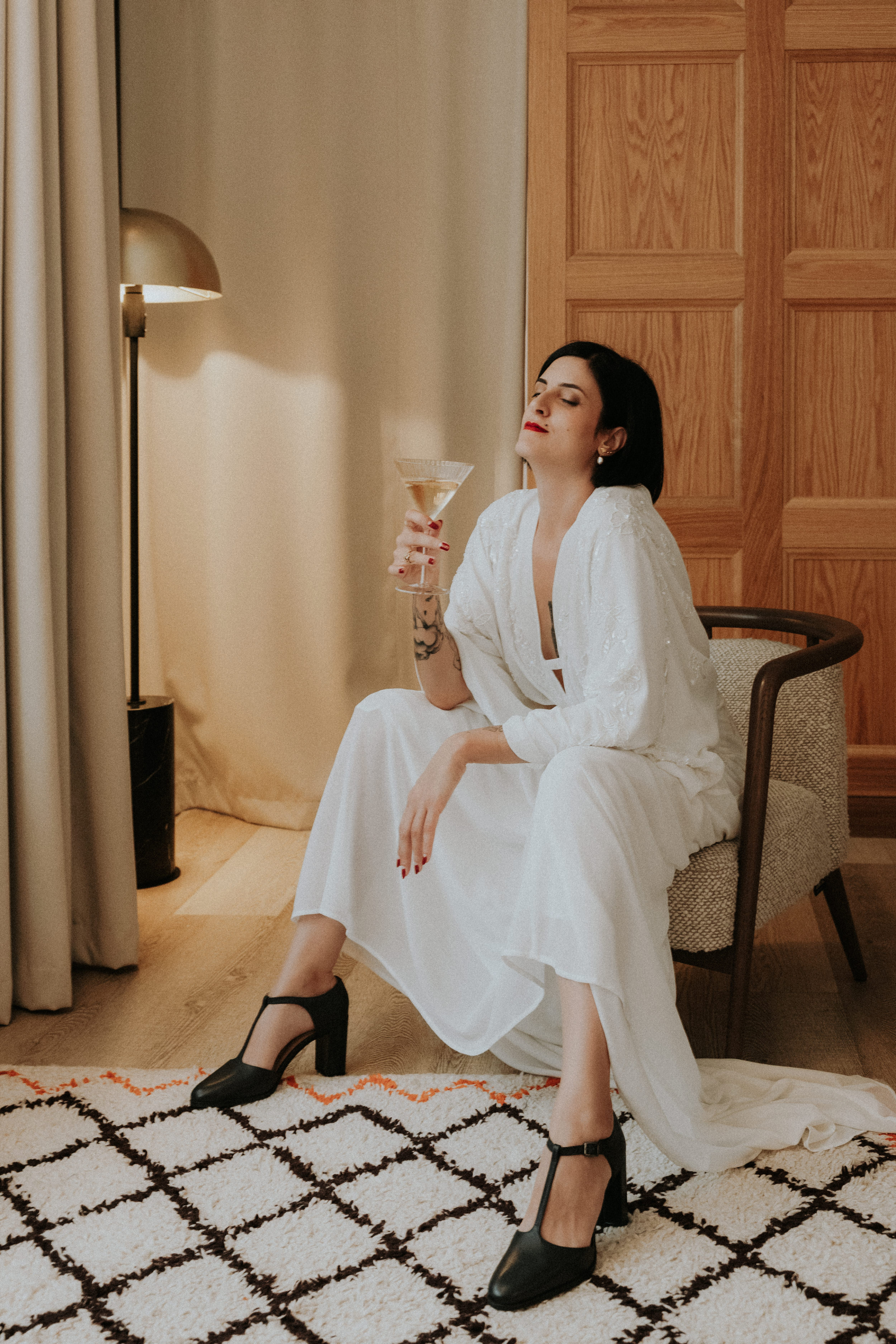 Bride in an ASOS wedding dress and T-bar black block heel shoes. She is sitting in a masculine pose with a glass of champagne. Bernadeta Kupiec Photography.