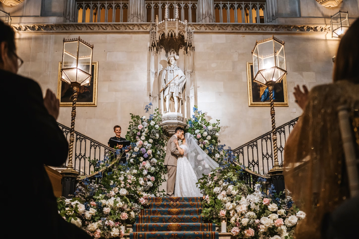 Bride and groom kissing at the top of a staircase at the end of their wedding ceremony. The staircase is lined with voluminous flowers finishing in a broken arch at the top. The celebrant stands nearby smiling. Vivienne May Weddings & Events - UK wedding planner. 
