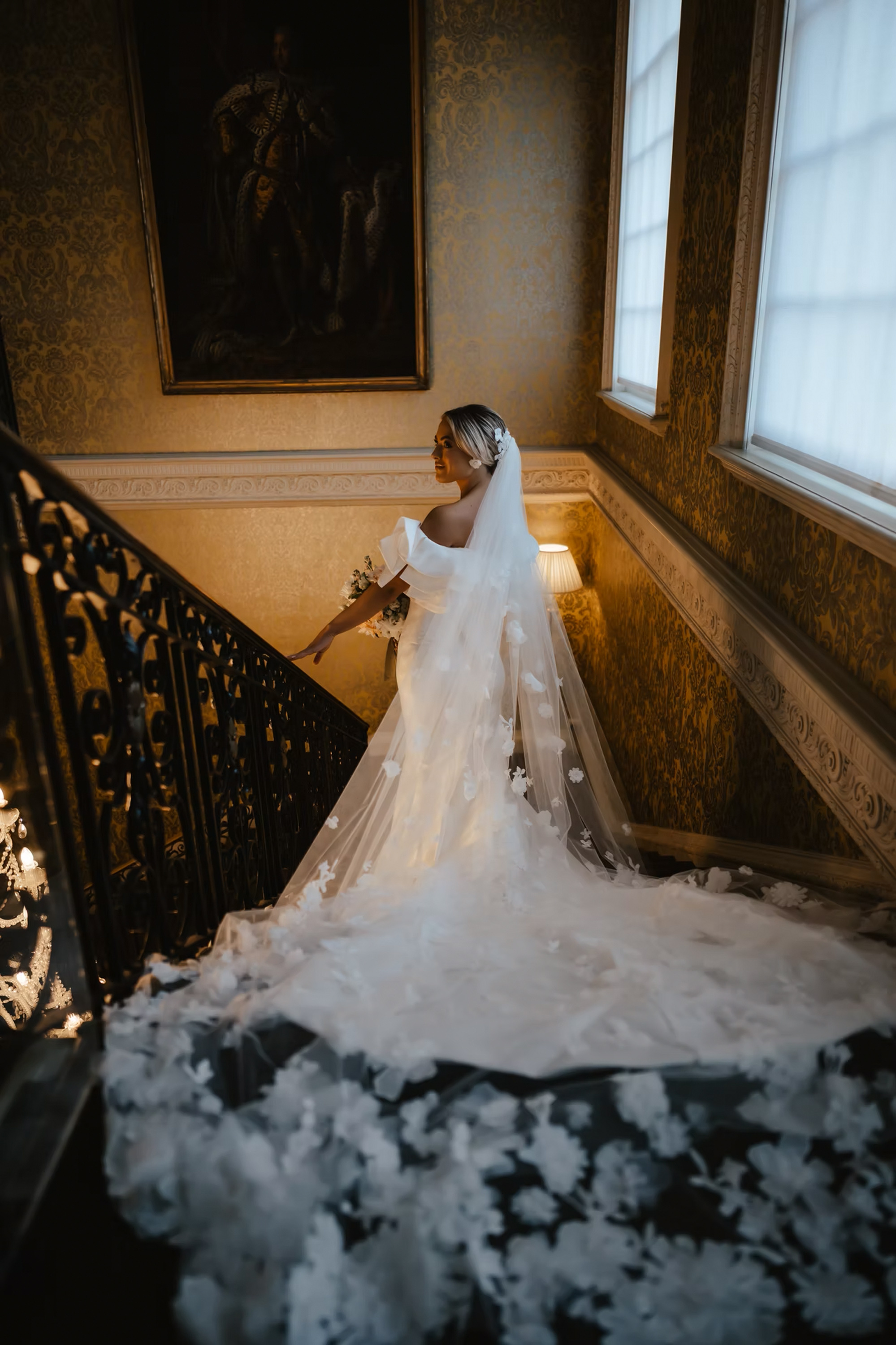 Bride standing on sweeping staircase in low light, a statement veil trailing behind her. Vivienne May Weddings & Events - UK wedding planner. 