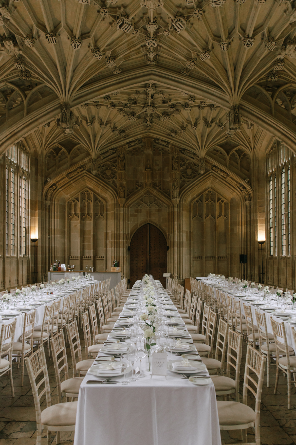A Classy & Modern City Wedding at the Bodleian Library With Jamaican-Guyanese Heritage 53 Stunning long table setup for a wedding reception in the Divinity School at the Bodleian Library, Oxford, featuring white linens, simple floral centrepieces, and a dramatic fan-vaulted gothic ceiling.