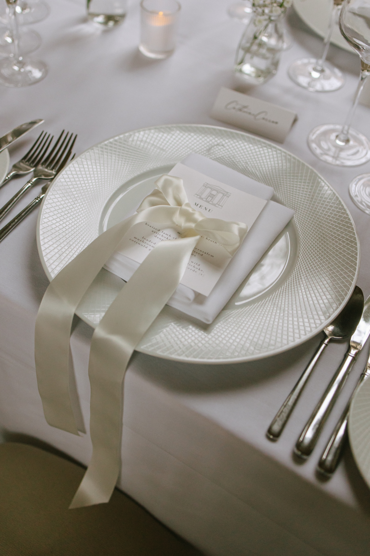A Classy & Modern City Wedding at the Bodleian Library With Jamaican-Guyanese Heritage 54 Close-up of a refined wedding place setting featuring a textured charger plate, personalised menu card tied with an ivory satin ribbon in a bow and minimalist calligraphy name card, styled on a crisp white tablecloth.