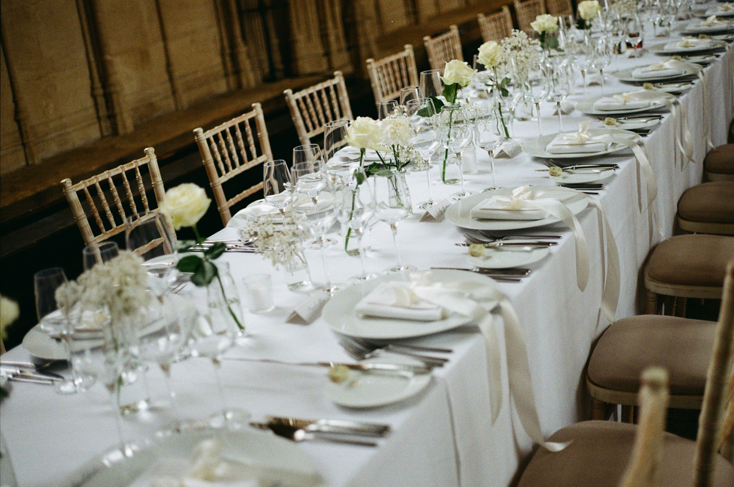 A Classy & Modern City Wedding at the Bodleian Library With Jamaican-Guyanese Heritage 29 Elegant long reception table set with crisp white linens, ivory ribbon-tied napkins, glassware and delicate white floral arrangements including roses and gypsophila, inside the historic Bodleian Library."