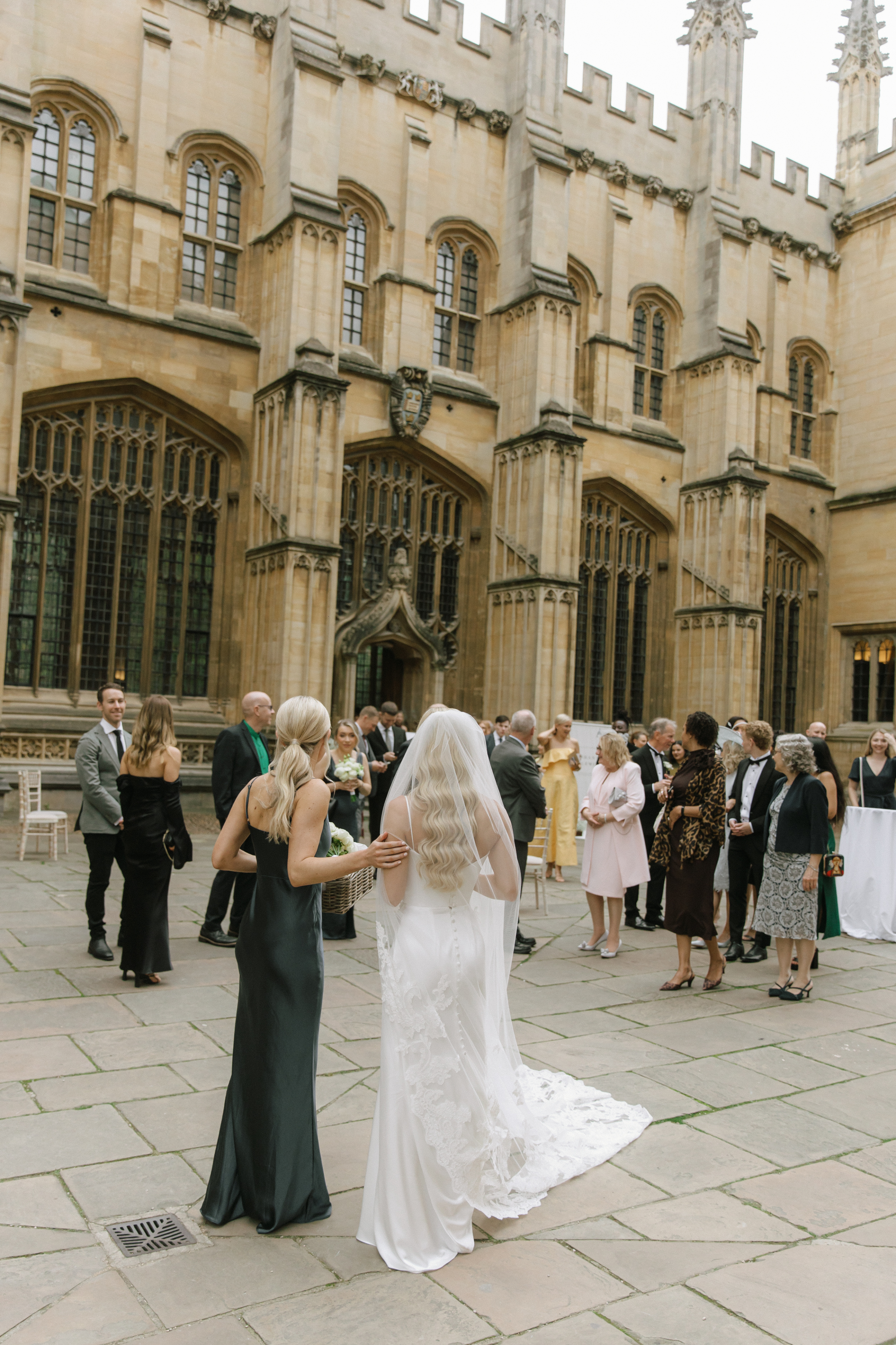 Guests mingle during the wedding drinks reception in the grand courtyard of the Bodleian Library, as the bride, with long flowing veil, is greeted by a bridesmaid in an emerald satin gown.