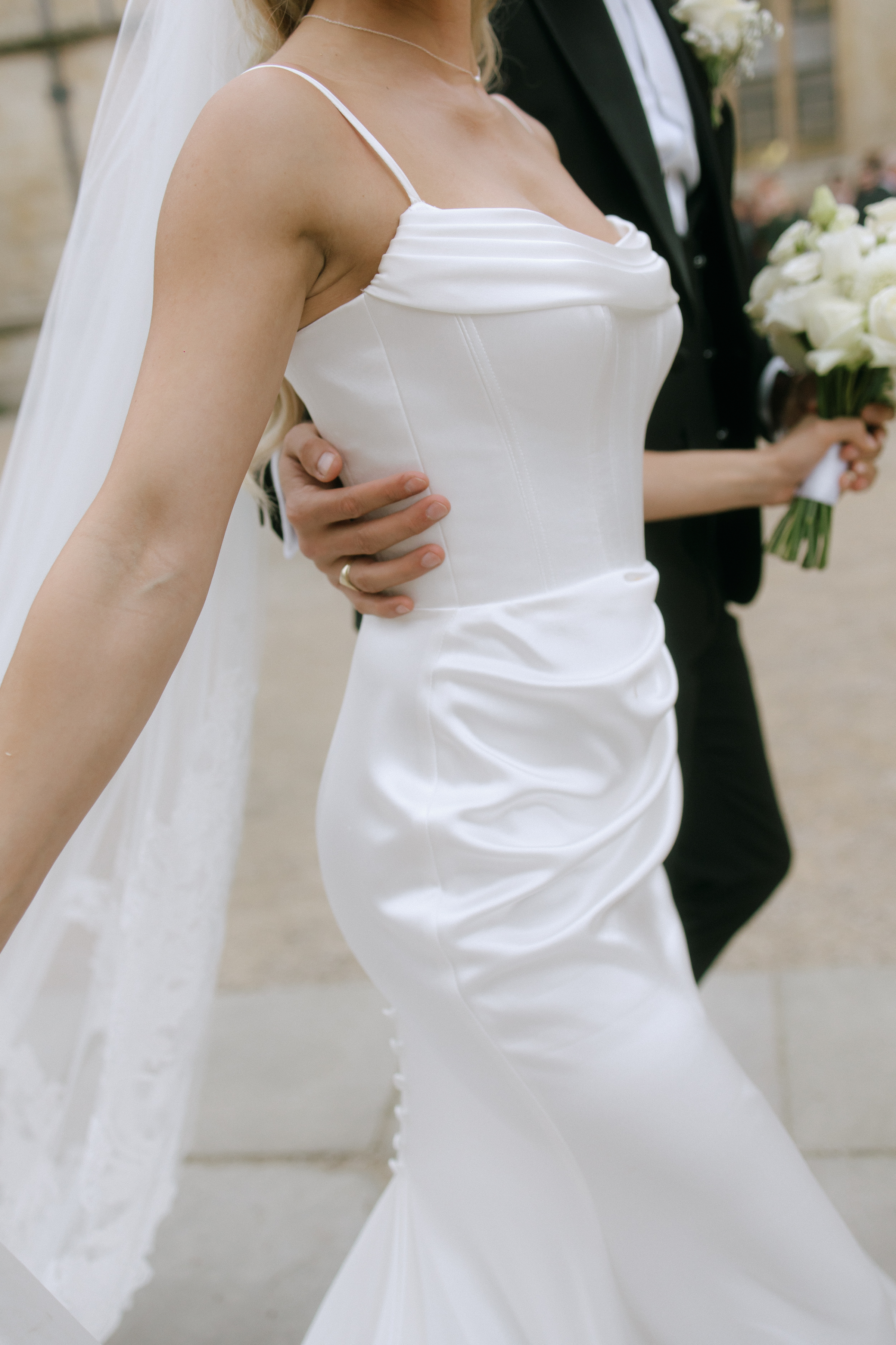 Close-up of the bride’s sleek silk wedding dress featuring a softly draped neckline and ruched hip detailing, as the groom holds her waist and carries a bouquet of ivory roses.