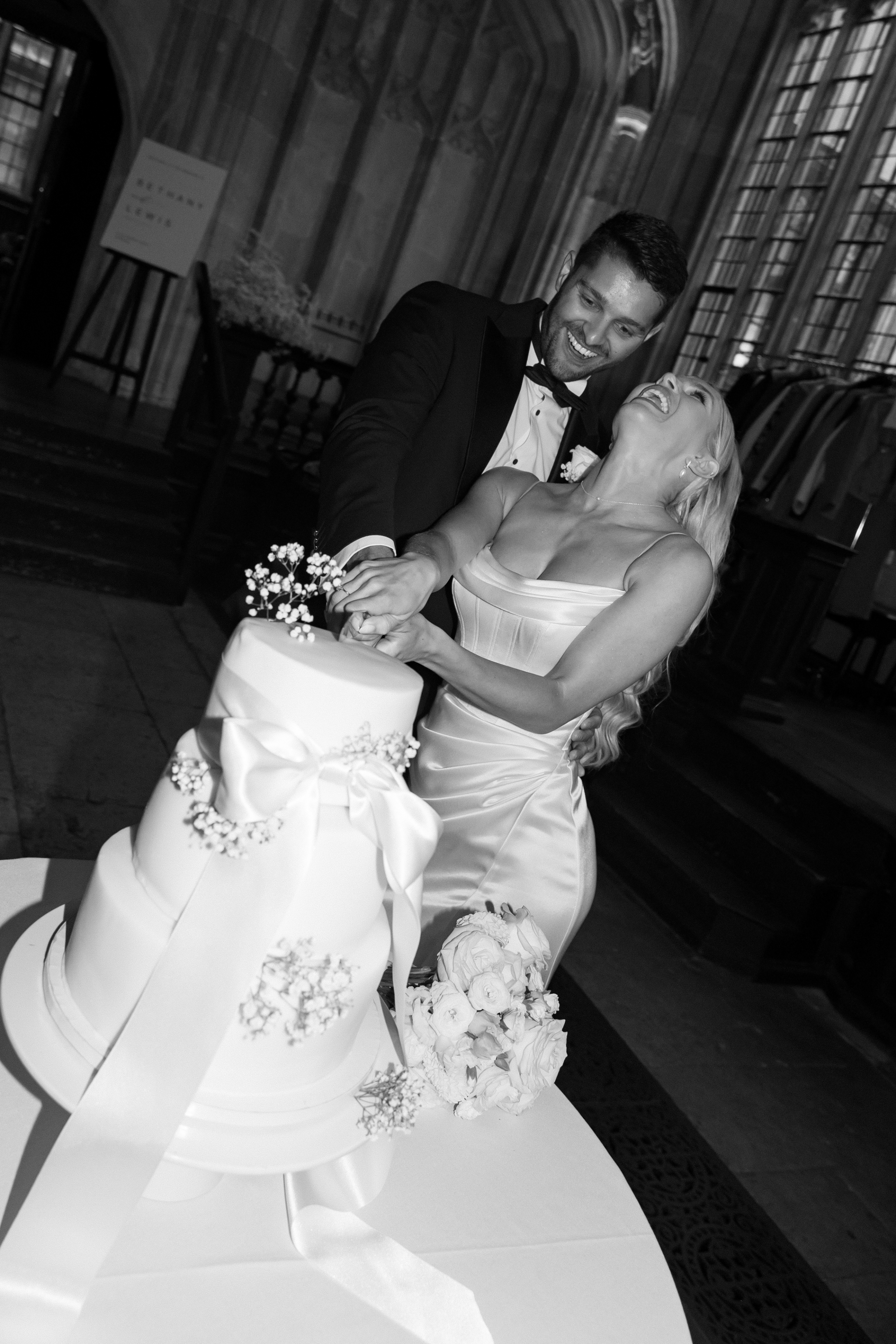 Black-and-white photo of the bride and groom laughing as they cut their white tiered wedding cake adorned with a satin ribbon and delicate baby’s breath.