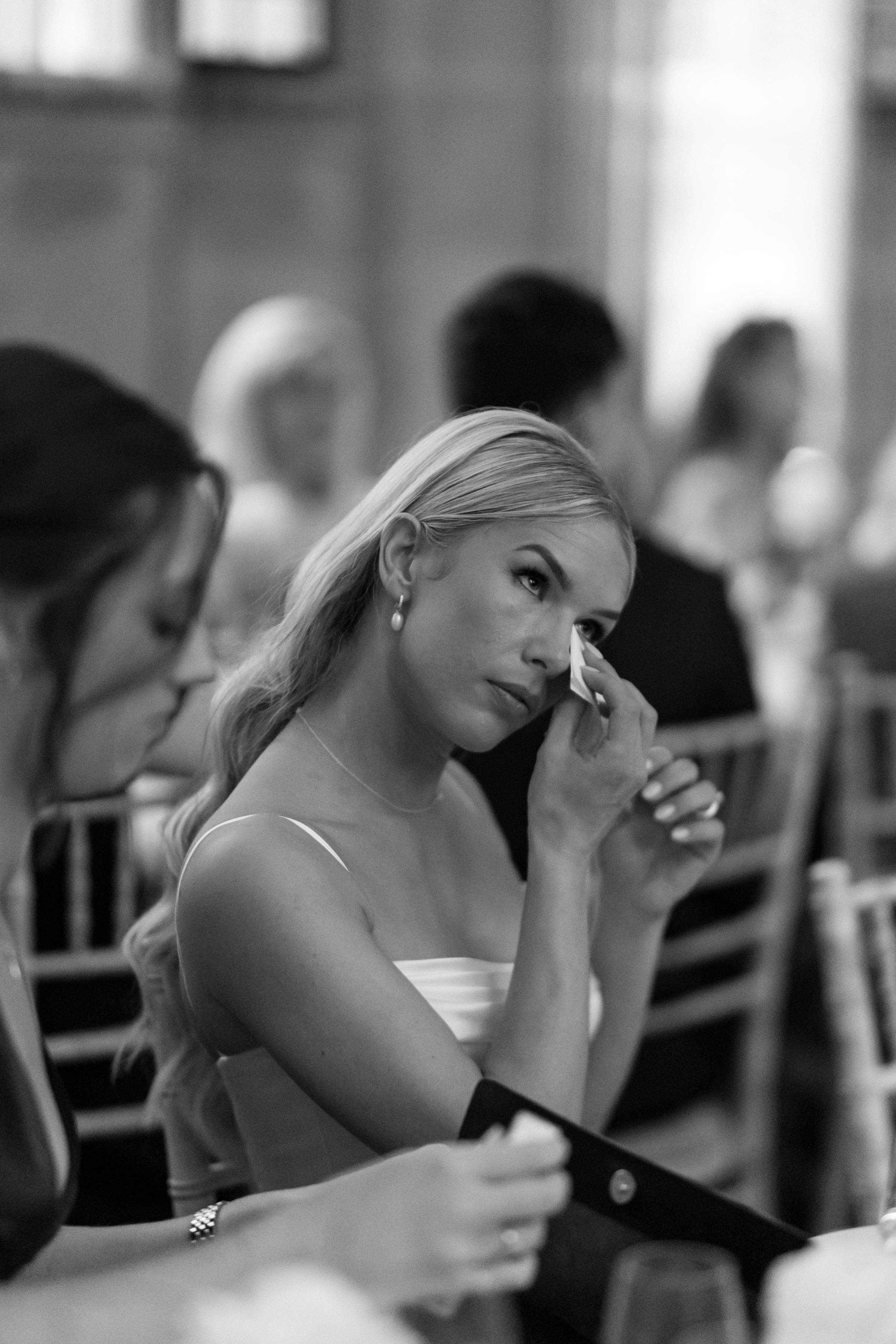 The bride delicately wipes away a tear while listening to a heartfelt speech, surrounded by guests at the candlelit wedding reception.