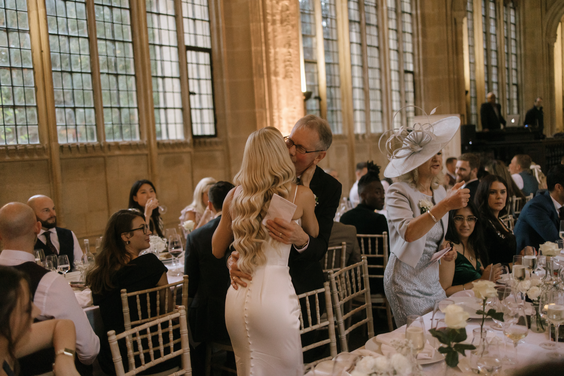 The bride shares a warm hug with her father during the wedding reception, captured mid-embrace beneath the grand windows of the Bodleian Library.