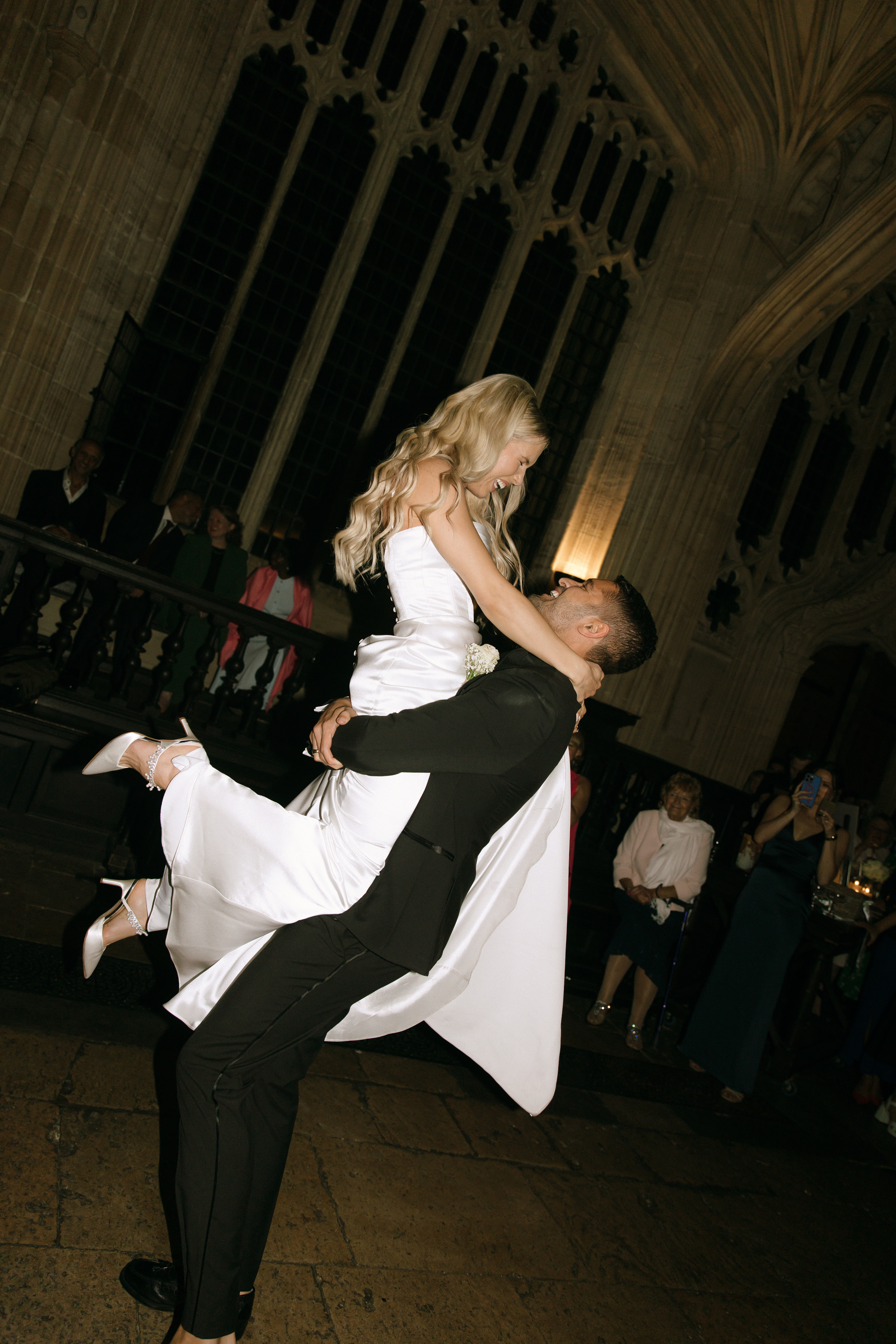 A Classy & Modern City Wedding at the Bodleian Library With Jamaican-Guyanese Heritage 64 The groom lifts the bride off her feet during their first dance, both laughing joyfully, as they celebrate surrounded by guests in the breathtaking interior of the Bodleian Library.