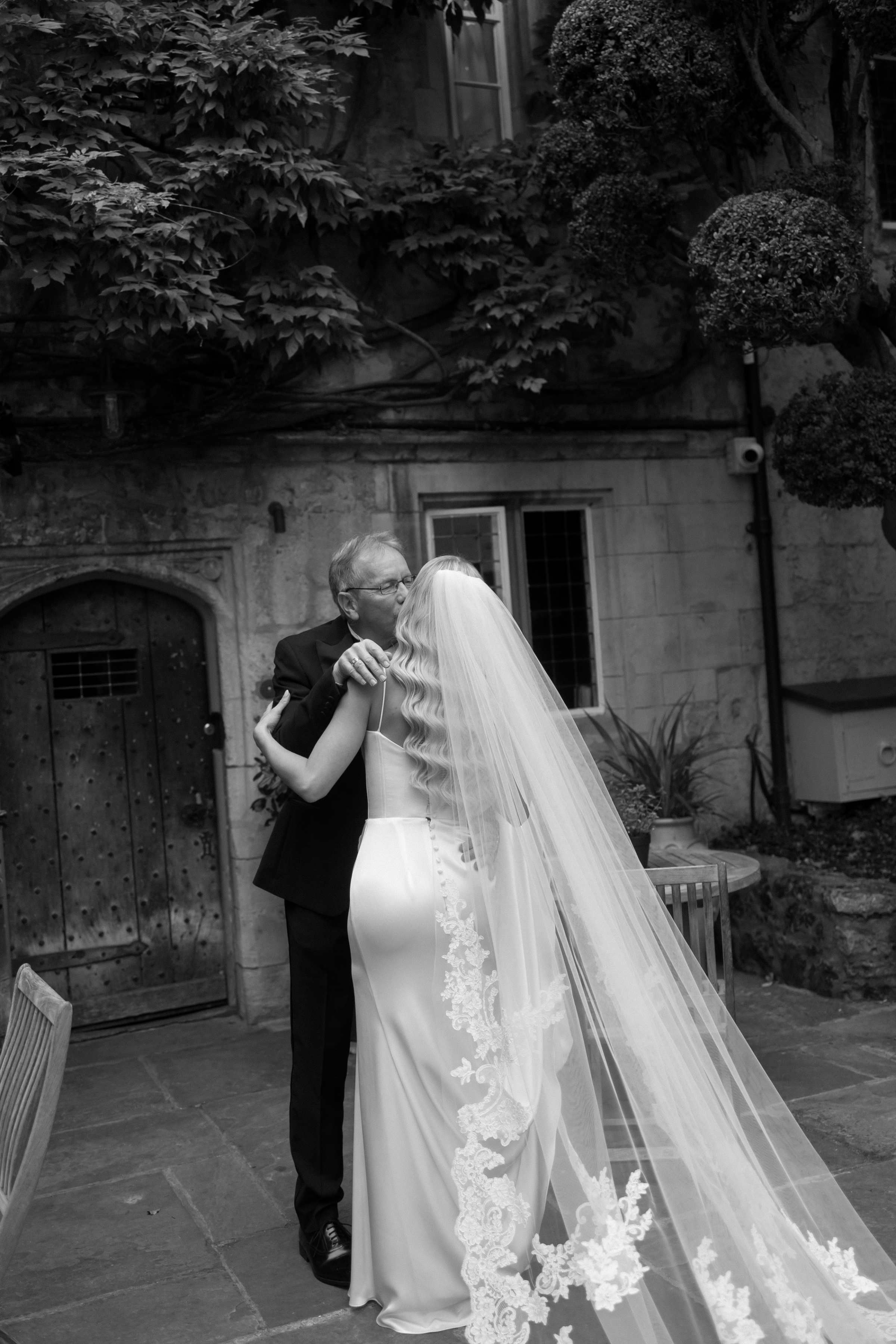 A Classy & Modern City Wedding at the Bodleian Library With Jamaican-Guyanese Heritage 34 Tender black-and-white moment as the bride shares an emotional hug and kiss with her father, her long embroidered veil flowing behind her in a courtyard setting.