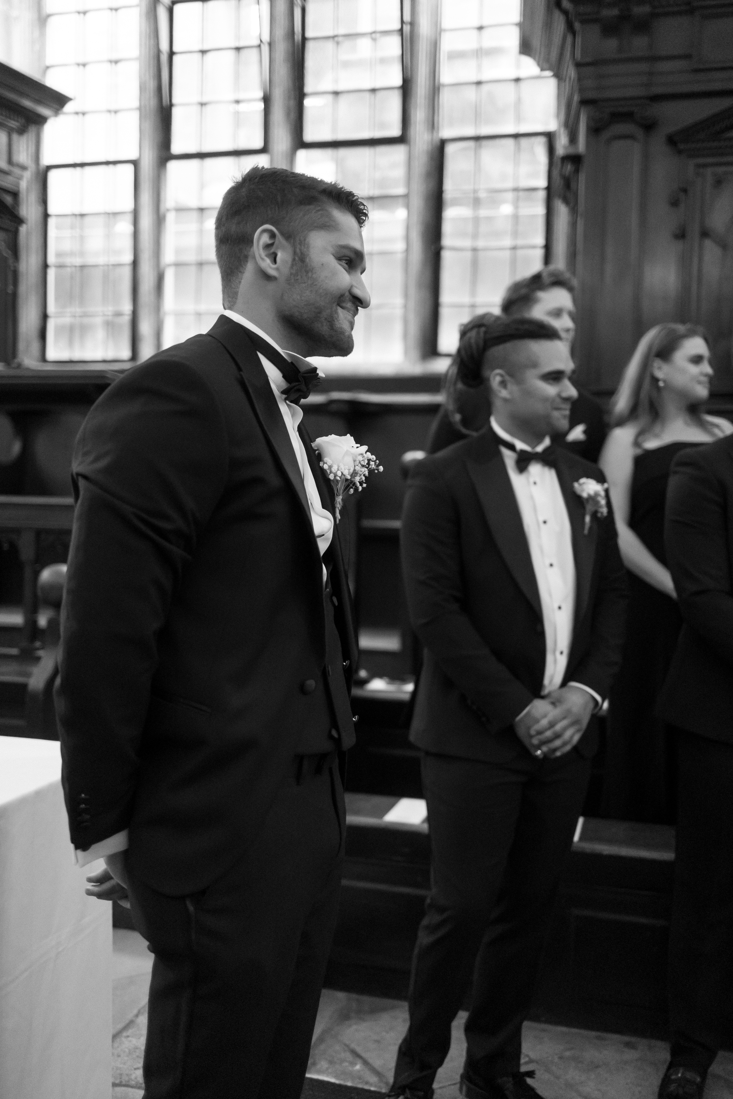 A Classy & Modern City Wedding at the Bodleian Library With Jamaican-Guyanese Heritage 35 Black-and-white image of the groom smiling in anticipation as he waits at the altar inside the historic Bodleian Library, flanked by groomsmen and surrounded by dark wooden panelling and arched windows.