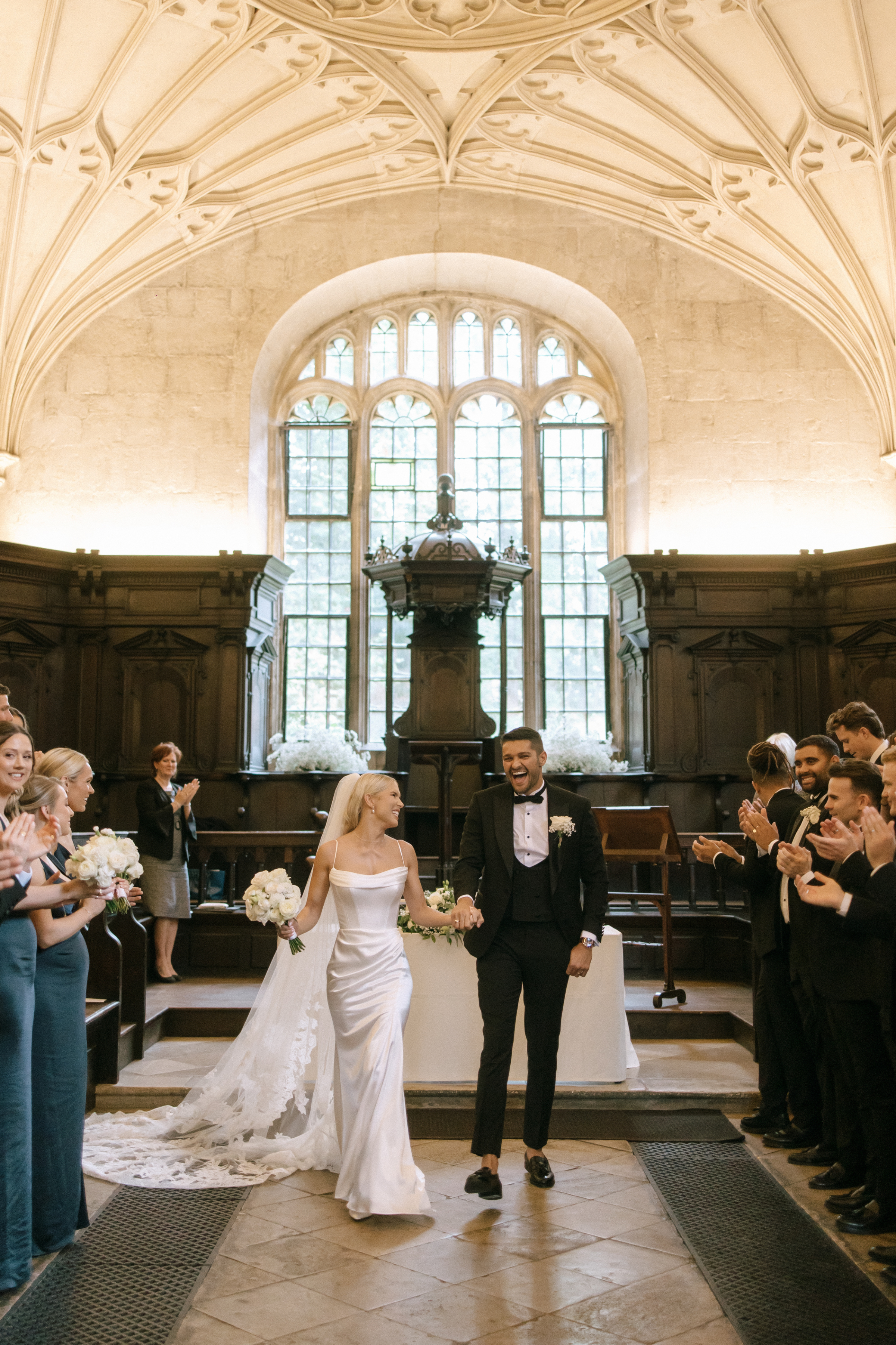 Bethany and Lewis walk back down the aisle hand-in-hand, smiling and surrounded by clapping guests in the Divinity School at the Bodleian Library, with floral displays and historic architecture behind them.
