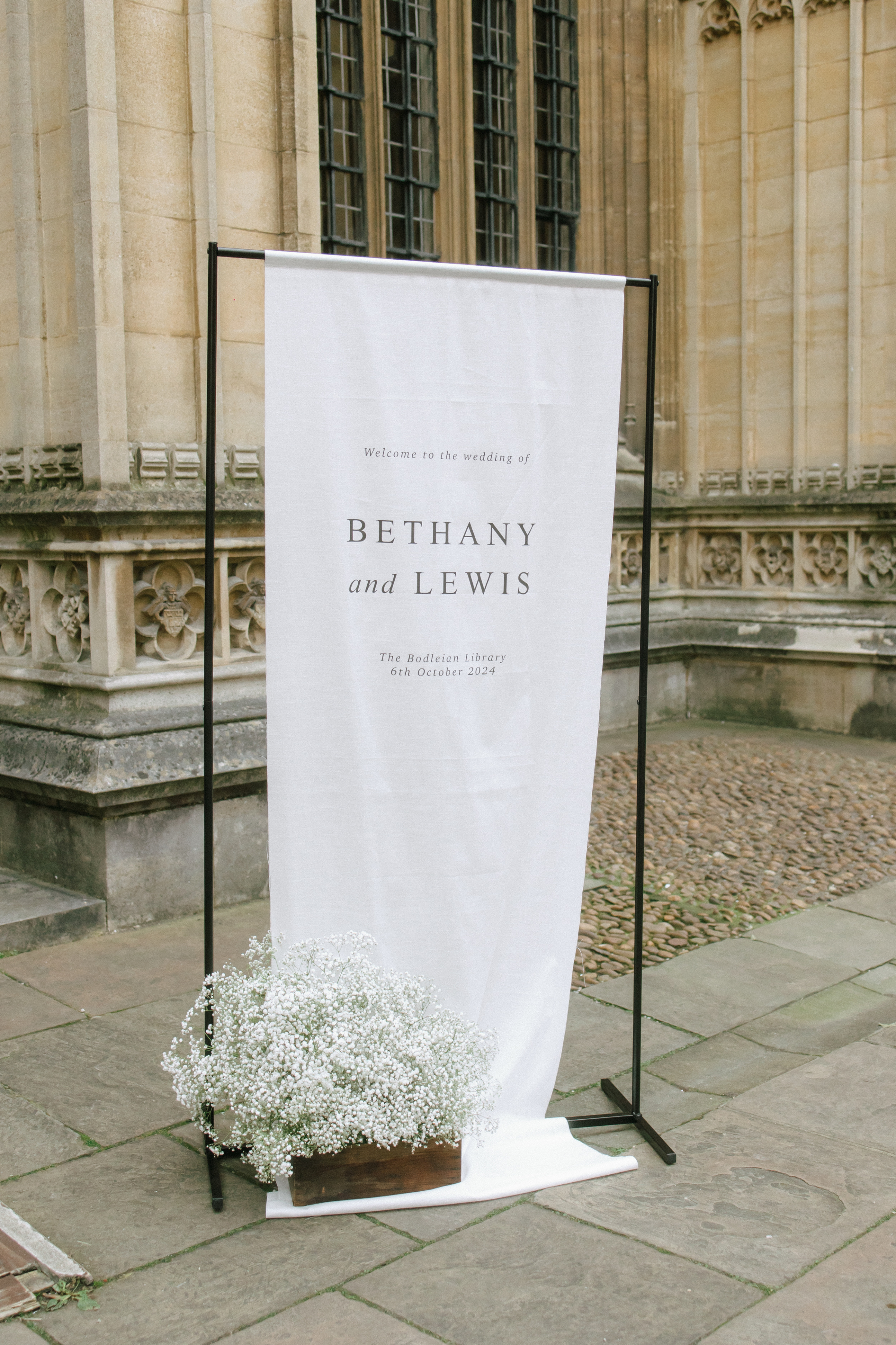 Modern white linen wedding welcome banner reading 'Bethany and Lewis, The Bodleian Library, 6th October 2024' displayed outside the historic venue, paired with soft white baby’s breath flowers.