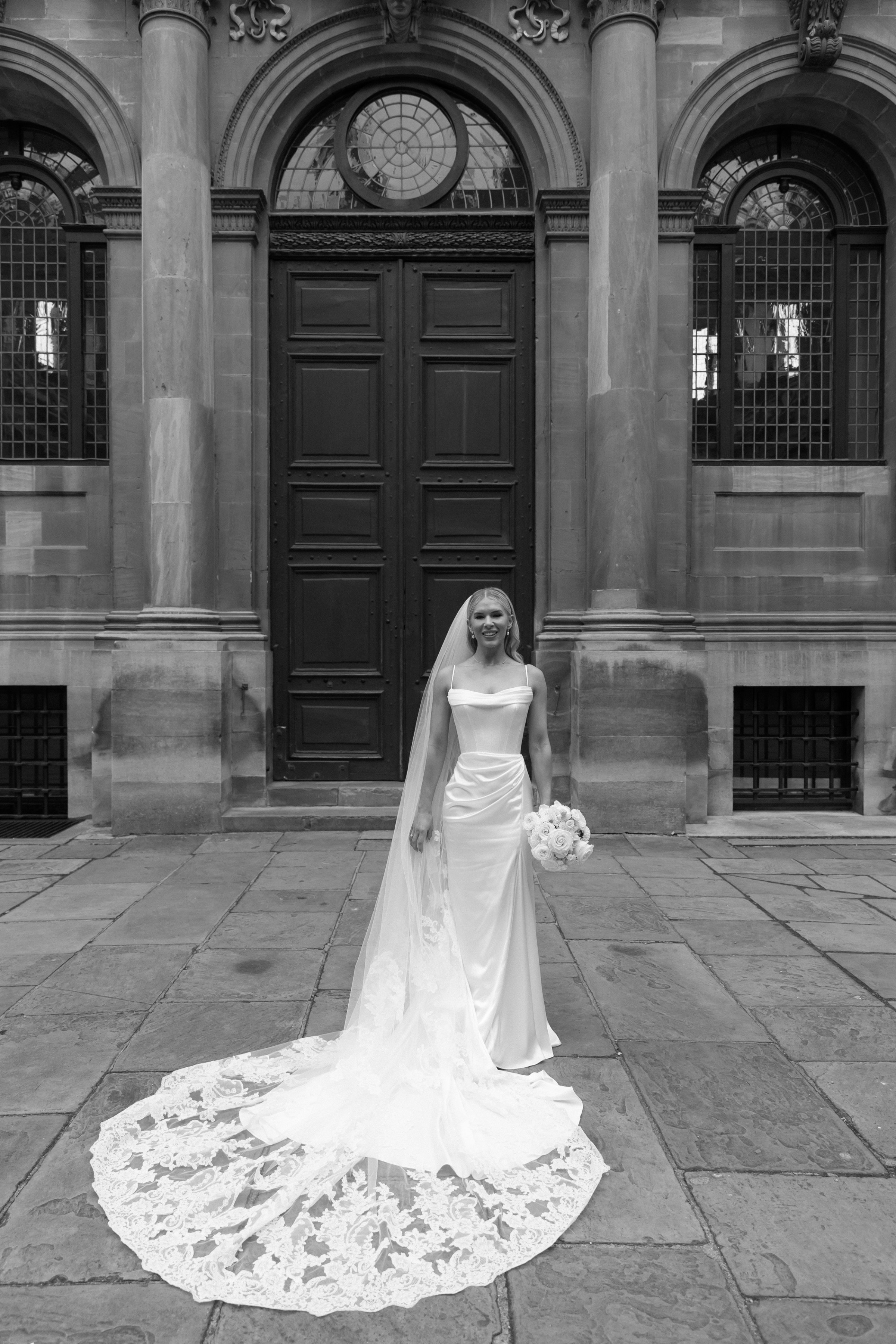 Black-and-white full-length image of the bride standing alone in front of grand arched doors, wearing a structured satin gown and cathedral-length embroidered veil fanned out in a perfect circle behind her.